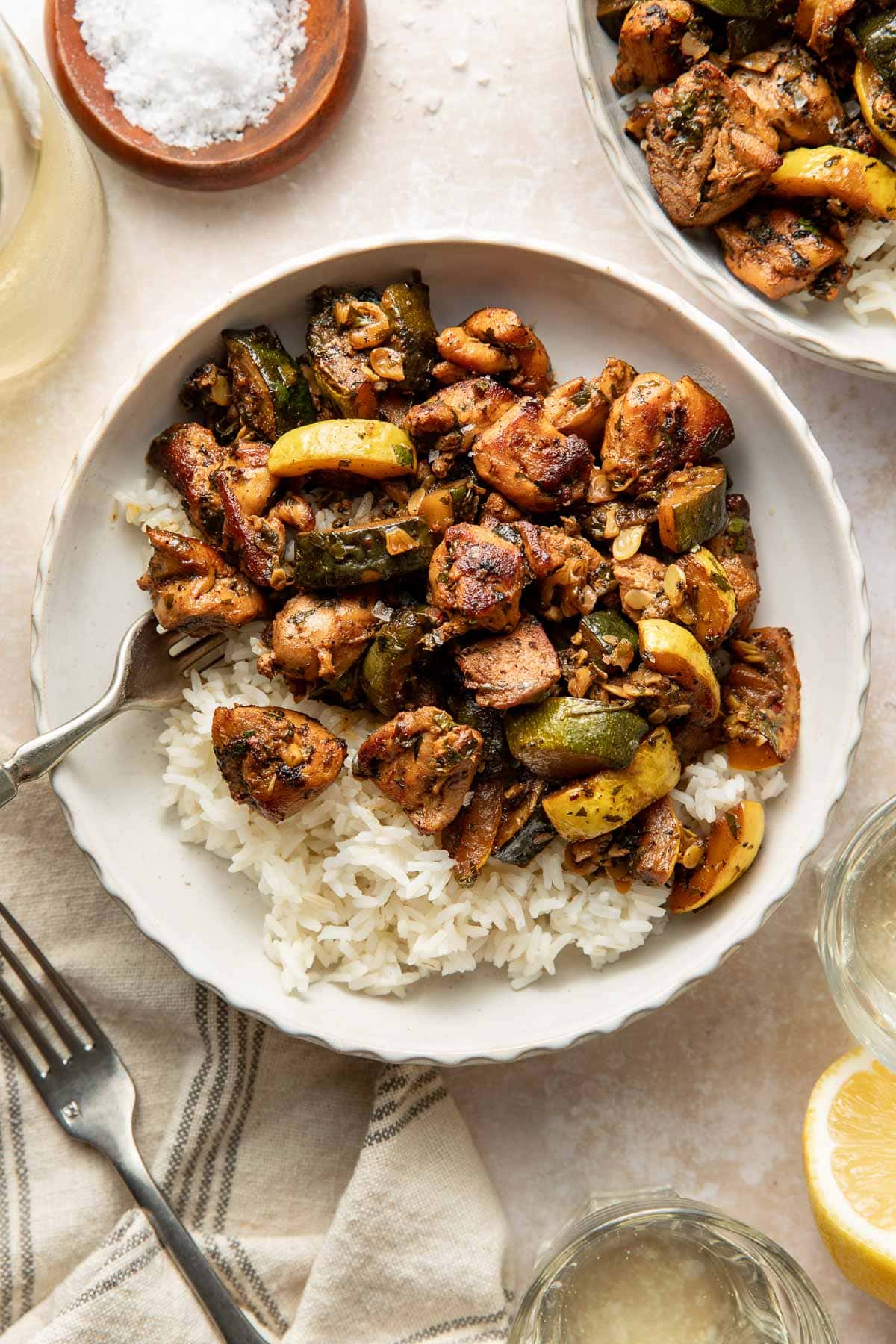 A white bowl filled with white rice topped with sautéed chicken pieces, zucchini, and yellow squash. A fork rests on the bowl, and glasses, a lemon slice, and a dish of salt are nearby.