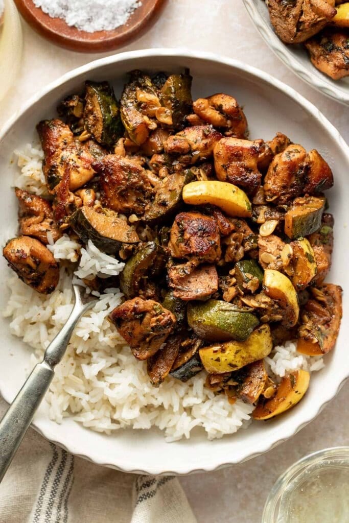 A bowl of white rice topped with sautéed chicken, zucchini, and yellow squash, seasoned with herbs and spices. A fork rests in the bowl, ready for eating.