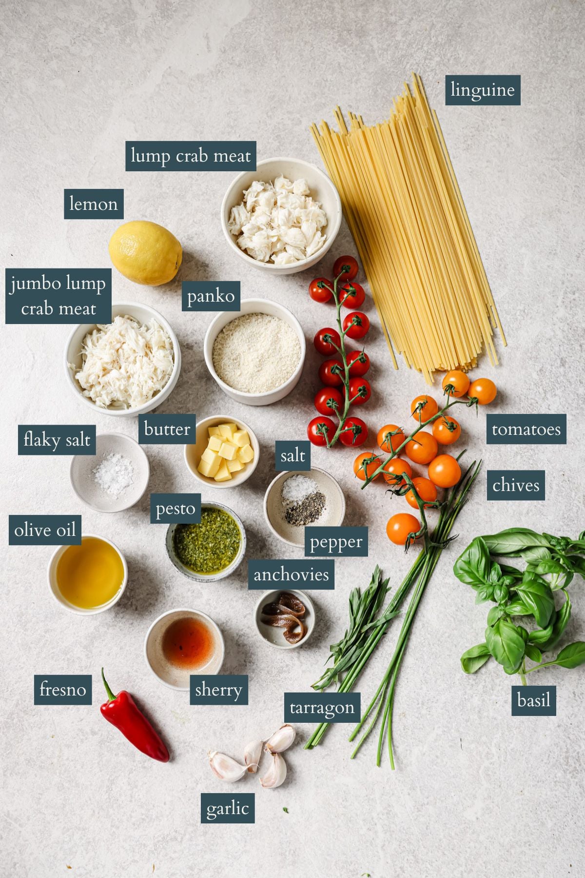 Overhead view of labeled ingredients for a pasta dish, including linguine, lump crab meat, lemon, panko, tomatoes, chives, butter, pesto, salt, pepper, anchovies, olive oil, tarragon, sherry, basil, garlic, and a Fresno chili.