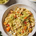 A bowl of linguine pasta with shredded chicken, roasted cherry tomatoes, and green herb breadcrumbs, served with a fork. Other bowls with breadcrumbs and pasta are in the background.