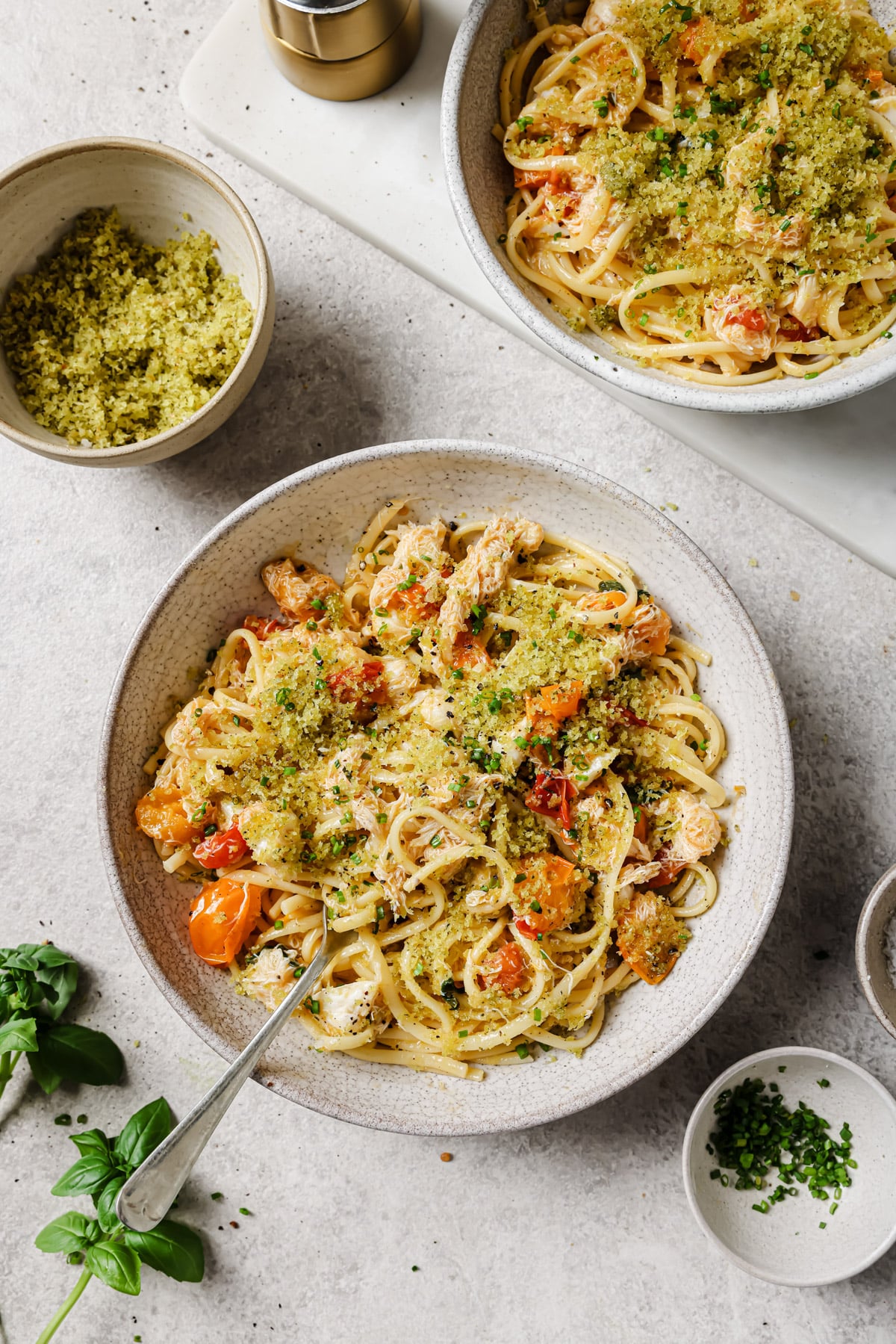 Two bowls of pasta topped with breadcrumbs, herbs, and pieces of chicken and cherry tomatoes are on a light surface. A small bowl of breadcrumbs, a dish of chopped herbs, and fresh basil leaves are nearby.