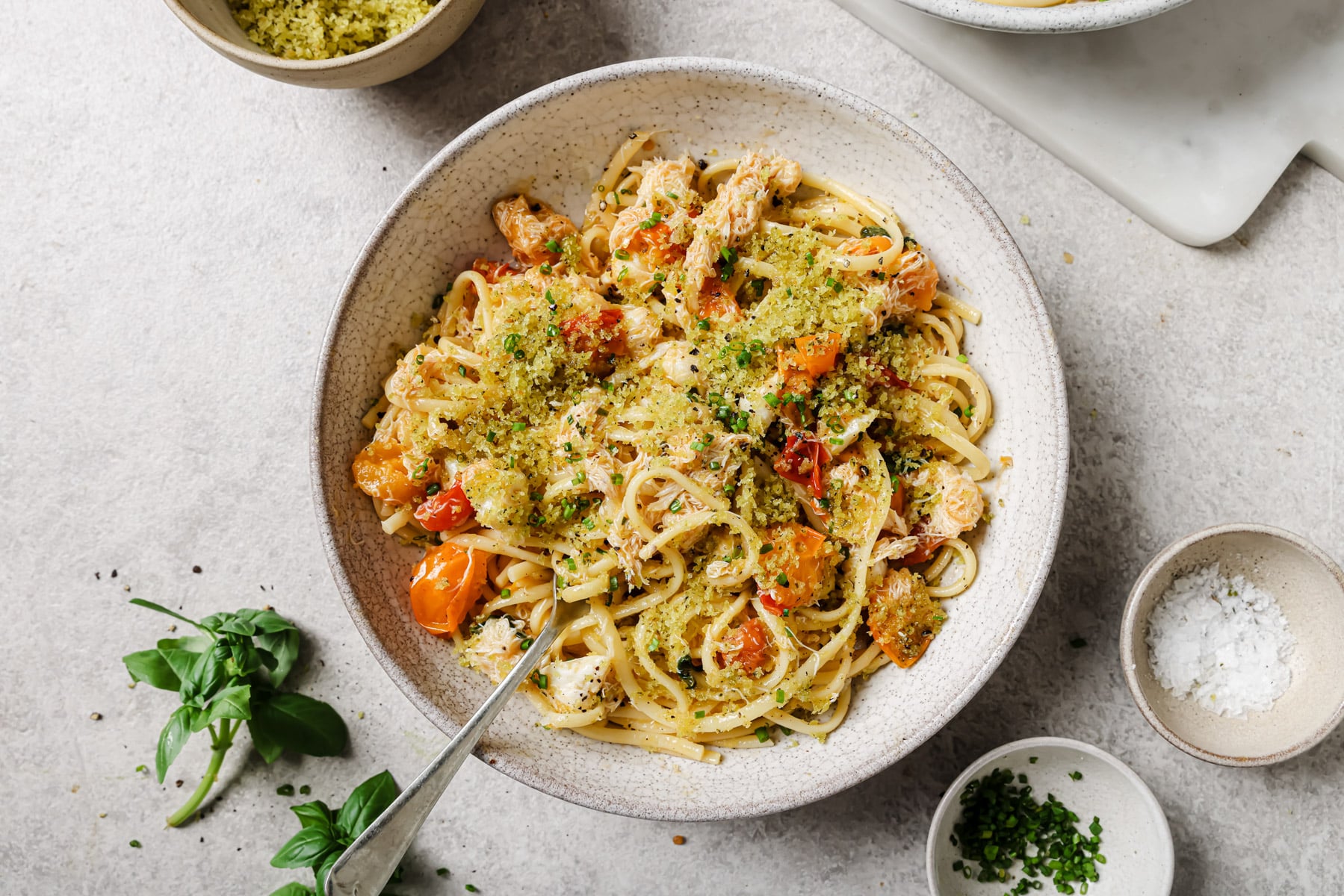 A bowl of linguine pasta topped with shredded chicken, roasted cherry tomatoes, green herbs, and breadcrumbs, with a fork in the bowl. Fresh basil, chopped herbs, and salt are arranged nearby on the table.