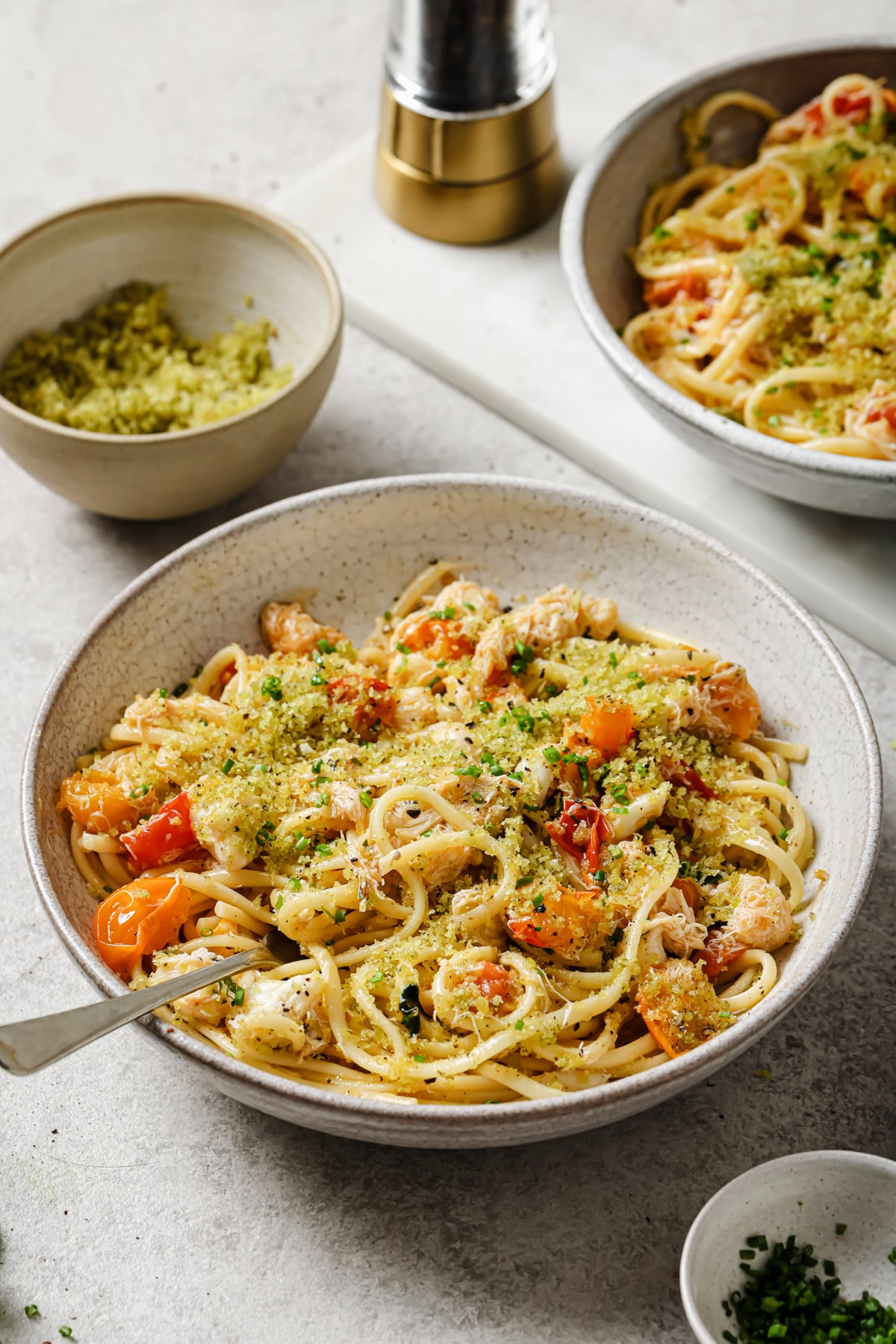 A bowl of pasta with cherry tomatoes, pesto, grated cheese, and fresh herbs sits on a table next to a small bowl of pesto, a pepper grinder, and another bowl of pasta in the background.