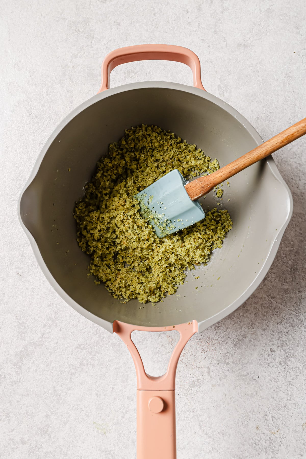 A beige pan with peach-colored handles contains finely chopped green vegetables being stirred with a blue spatula on a light gray surface.