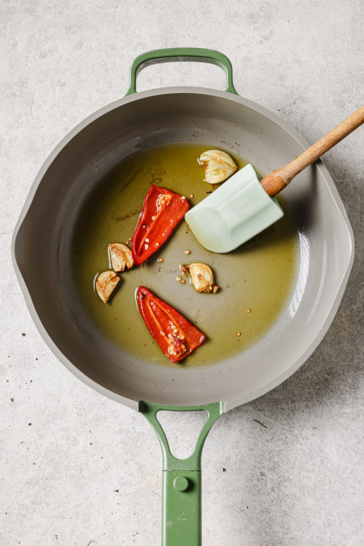 A gray skillet with a green handle contains olive oil, garlic cloves, and a sliced red chili pepper being stirred with a light green spatula on a light-colored surface.