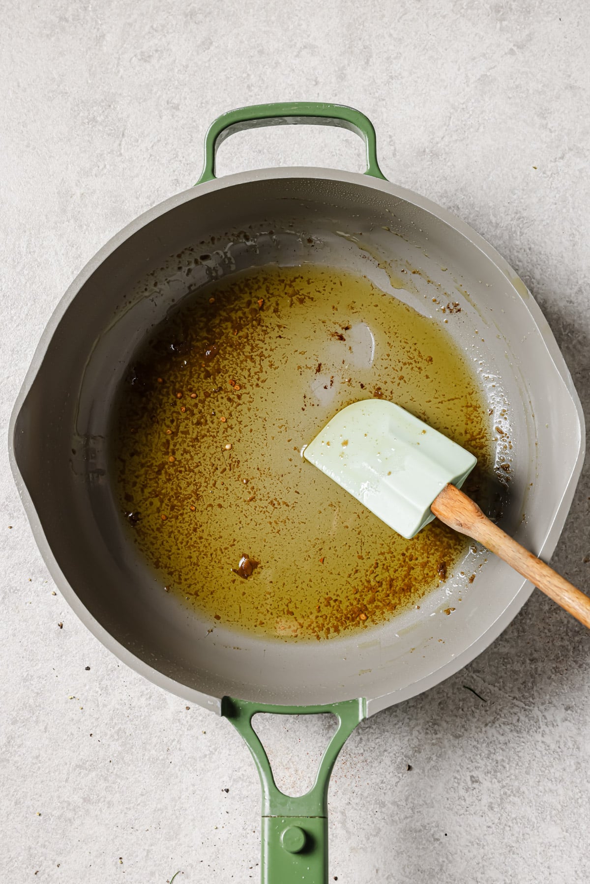 A gray skillet with green handles contains oil with spices. A light green spatula with a wooden handle rests in the oil.