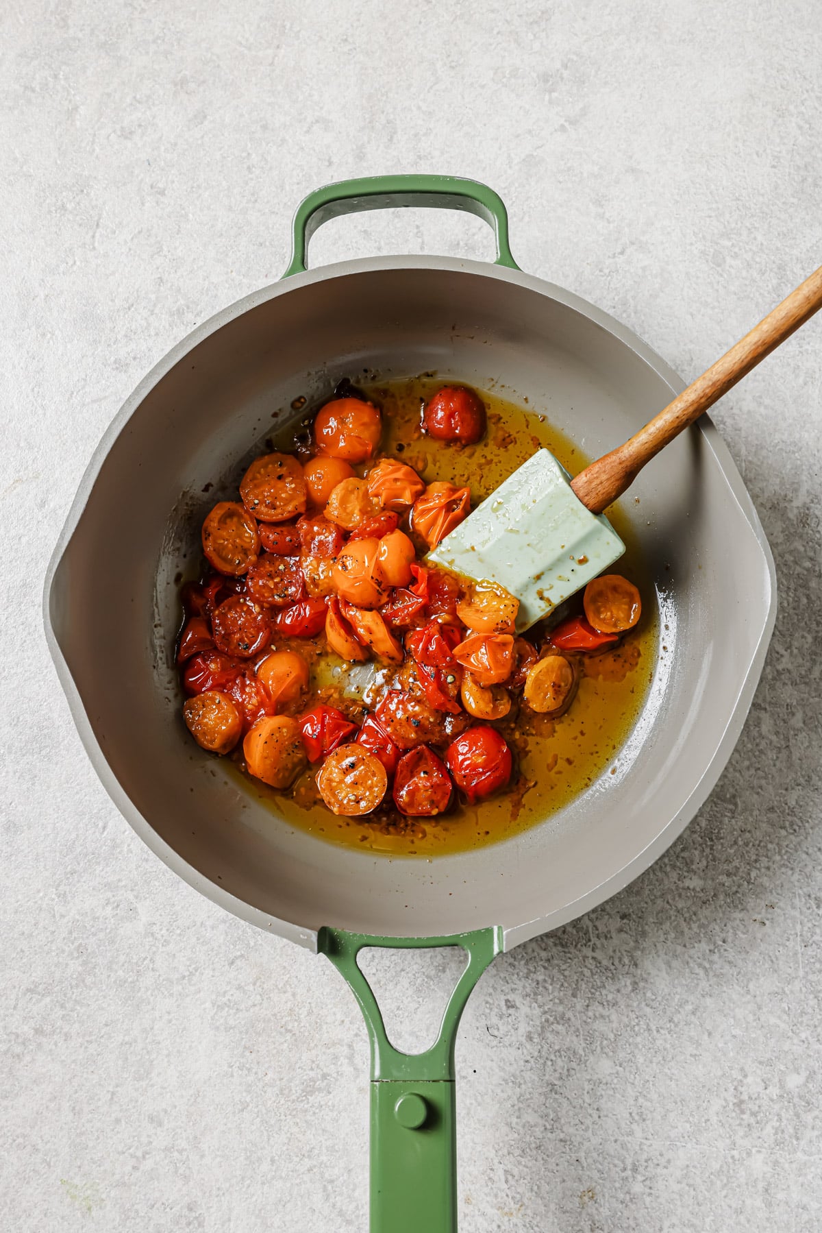 A green-handled skillet containing halved cherry tomatoes simmering in olive oil, being stirred with a light blue spatula on a light gray countertop.