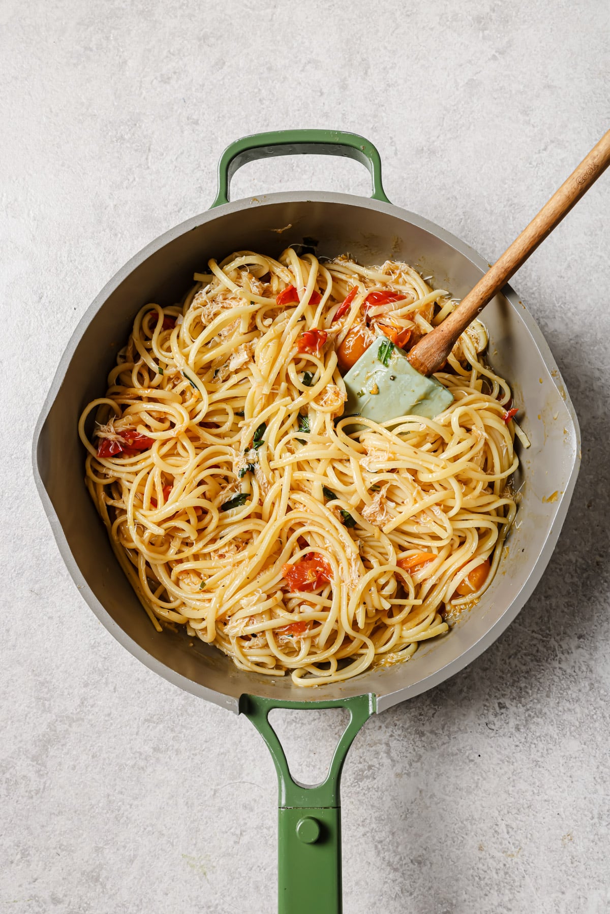 A green-handled pan filled with cooked spaghetti, mixed with sliced red peppers, herbs, and a creamy sauce. A spatula rests inside the pan, stirring the pasta. The surface beneath the pan is a light, textured countertop.
