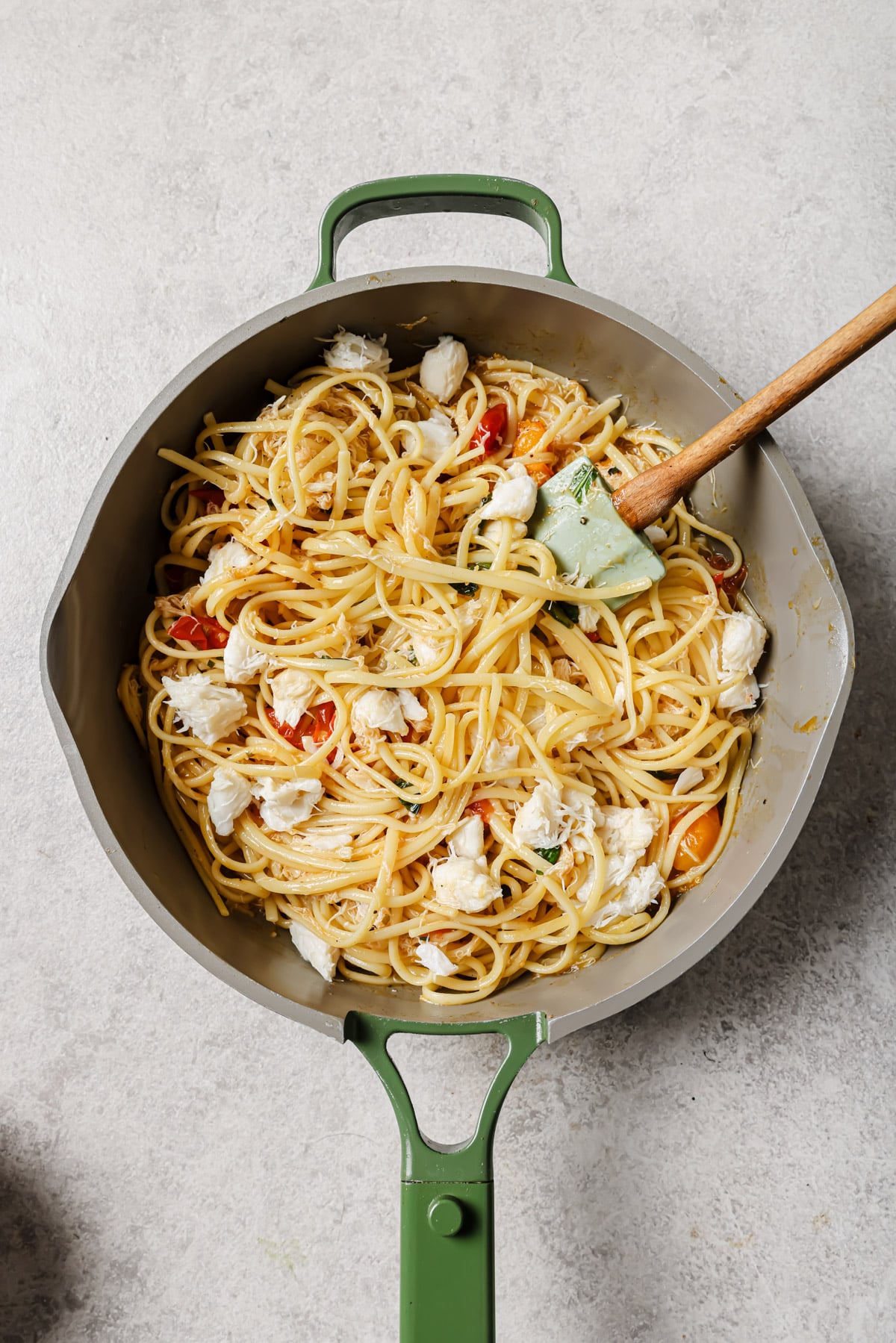 A green-handled skillet filled with spaghetti, cherry tomatoes, and chunks of white cheese or crab meat, with a green spatula resting inside. The skillet sits on a light-colored surface.