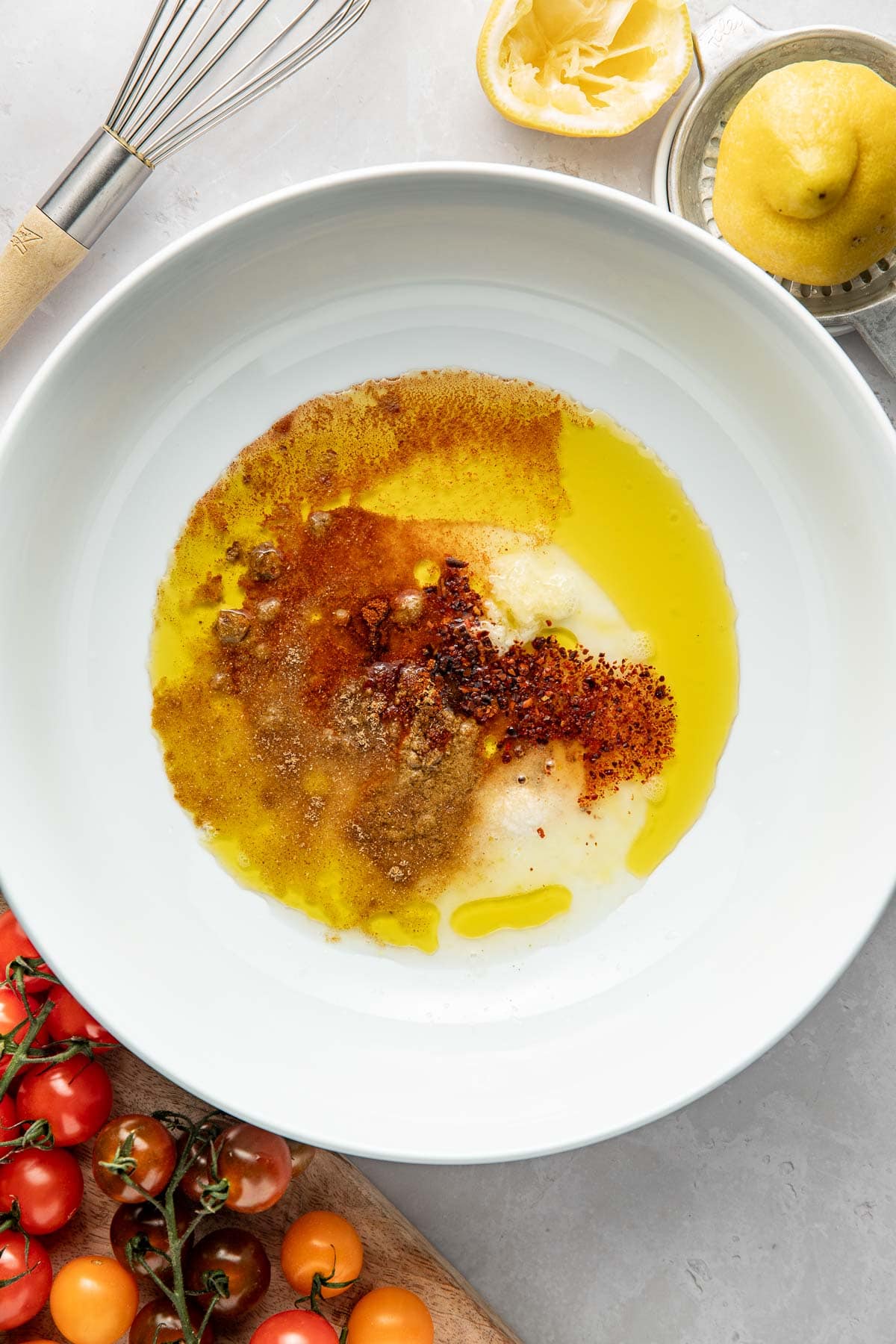 A white bowl contains olive oil, ground spices, and minced garlic, ready to be mixed. Surrounding the bowl are a whisk, a squeezed lemon, whole lemons, and a cutting board with red and yellow cherry tomatoes.