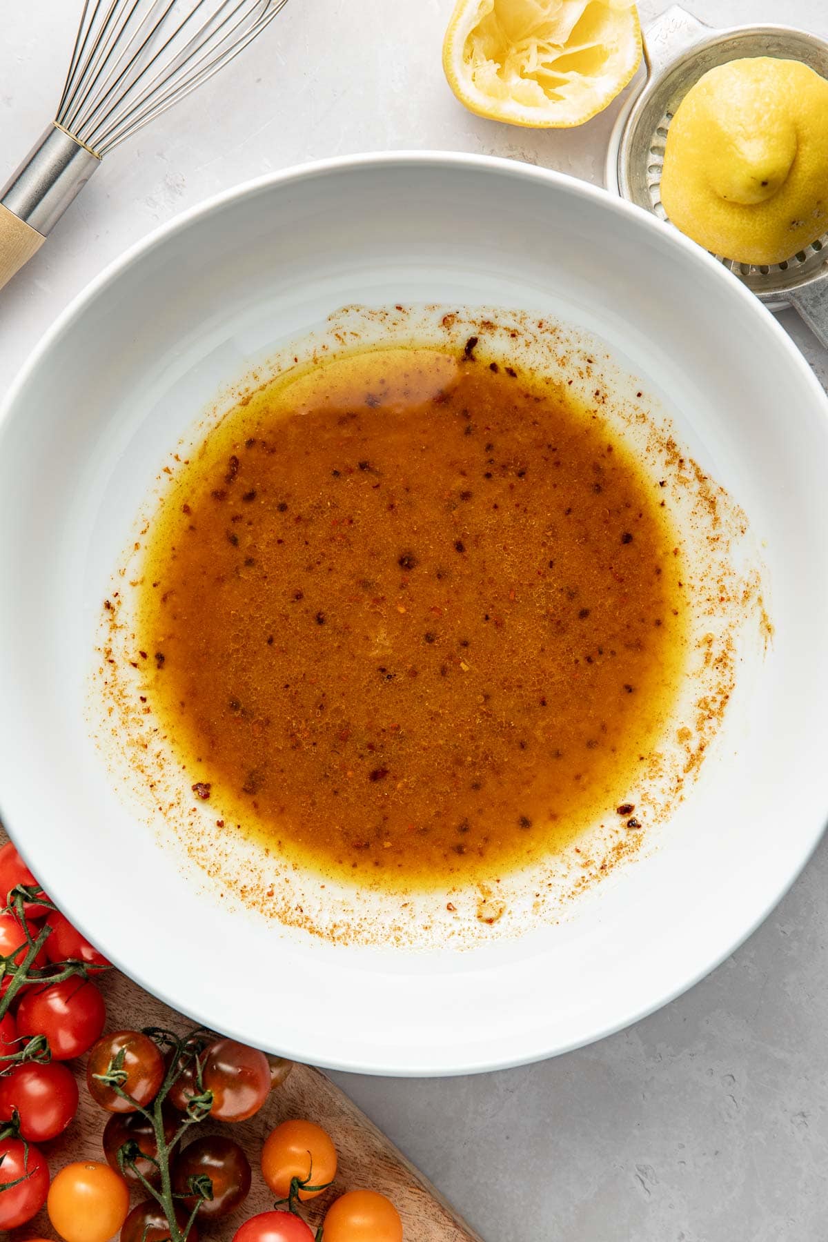 A white bowl containing a brown vinaigrette sits on a light surface, surrounded by a whisk, a lemon half, a citrus juicer, and a cutting board with assorted cherry tomatoes.