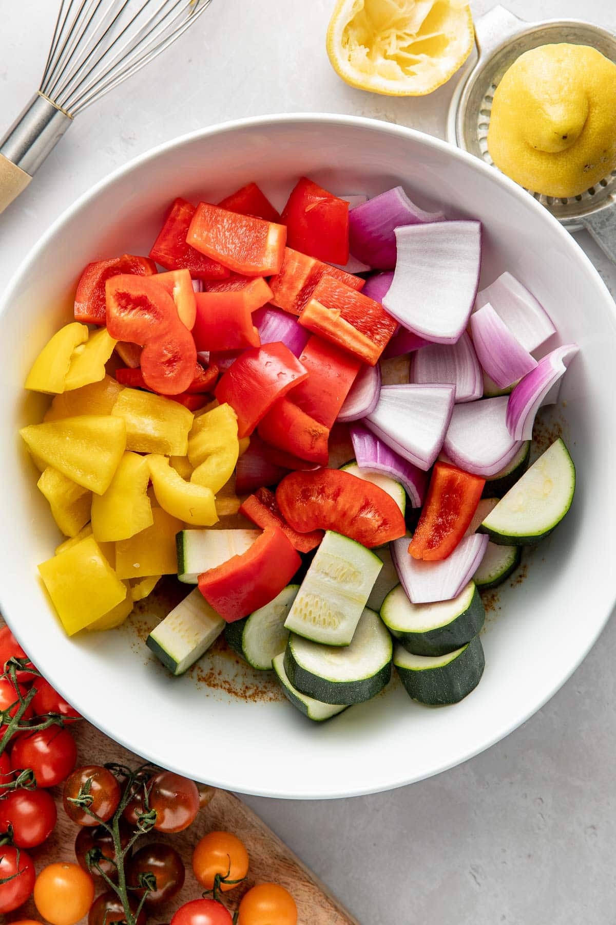 A white bowl filled with chopped yellow and red bell peppers, red onion, zucchini, and red tomatoes. A whisk, squeezed lemon, and fresh tomatoes on a cutting board are nearby on a light surface.