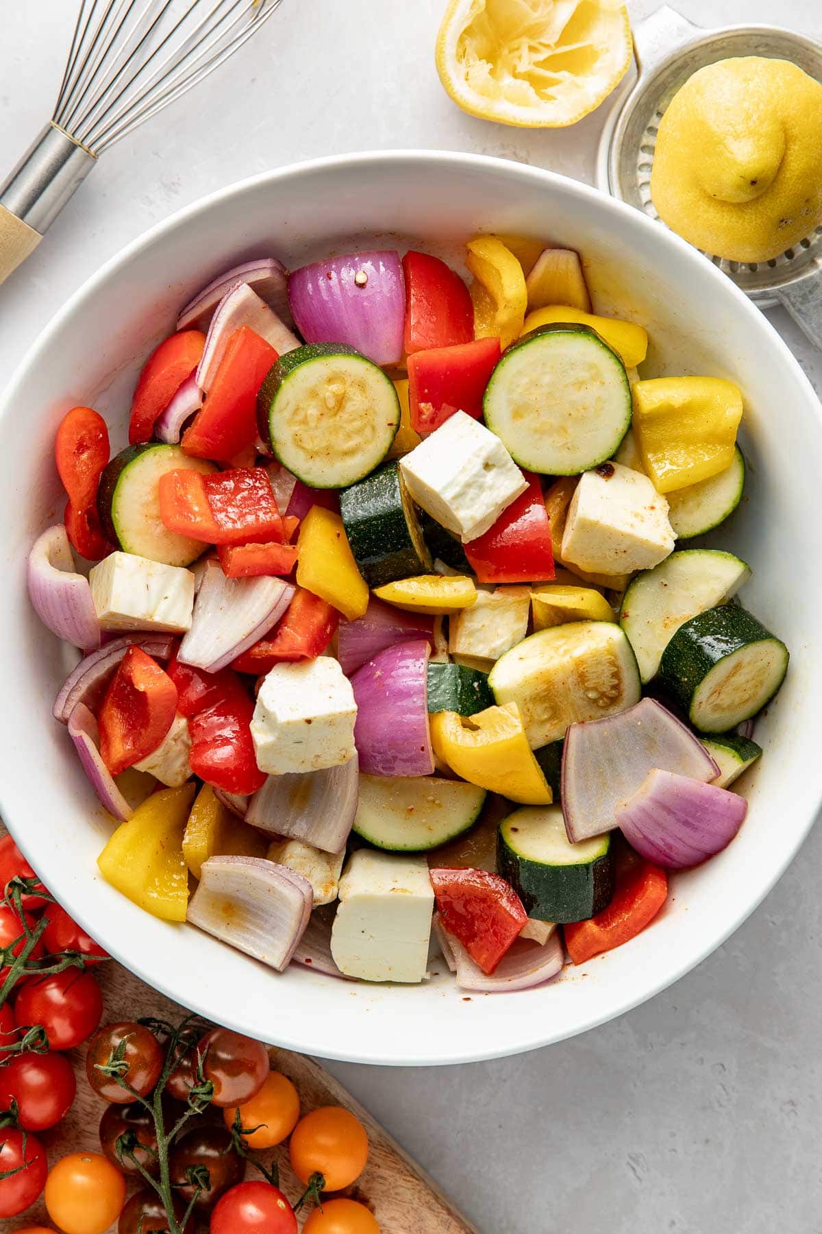 A white bowl filled with chopped raw vegetables, including zucchini, red and yellow bell peppers, red onion, tomatoes, and chunks of white cheese, surrounded by fresh lemons and cherry tomatoes on a light surface.