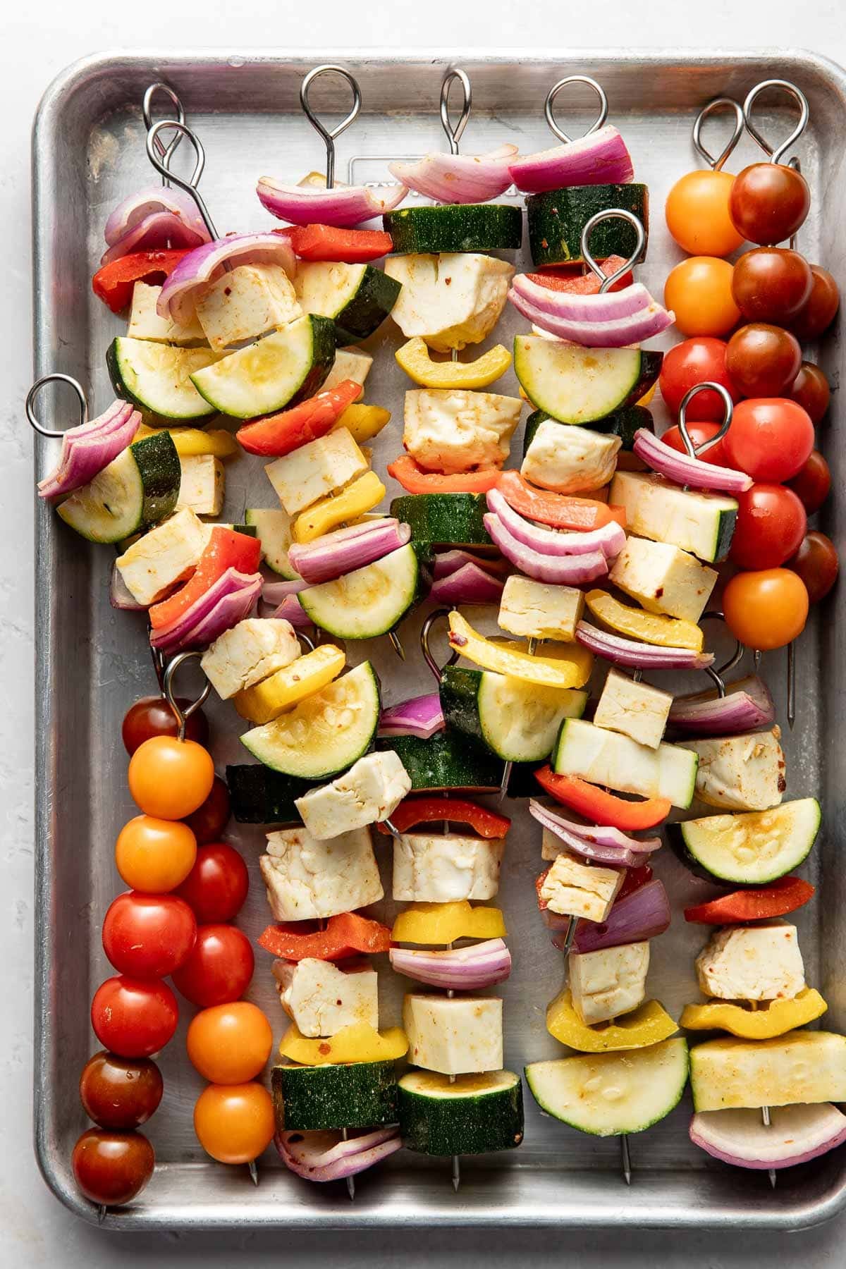 Skewers on a metal tray, loaded with cherry tomatoes, red onion, zucchini, yellow squash, red bell pepper, and cubes of tofu. The vegetables and tofu are arranged in colorful, alternating patterns.