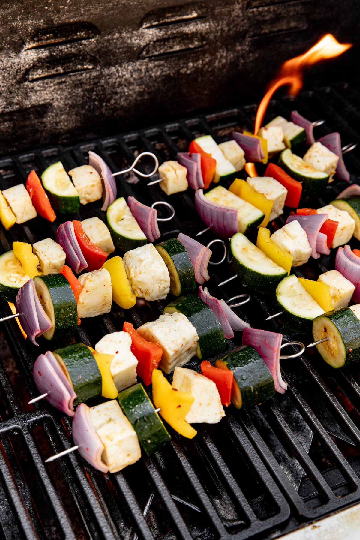 Colorful vegetable and tofu skewers with zucchini, bell peppers, red onion, and tofu grilling on a barbecue, with a flame visible in the background.