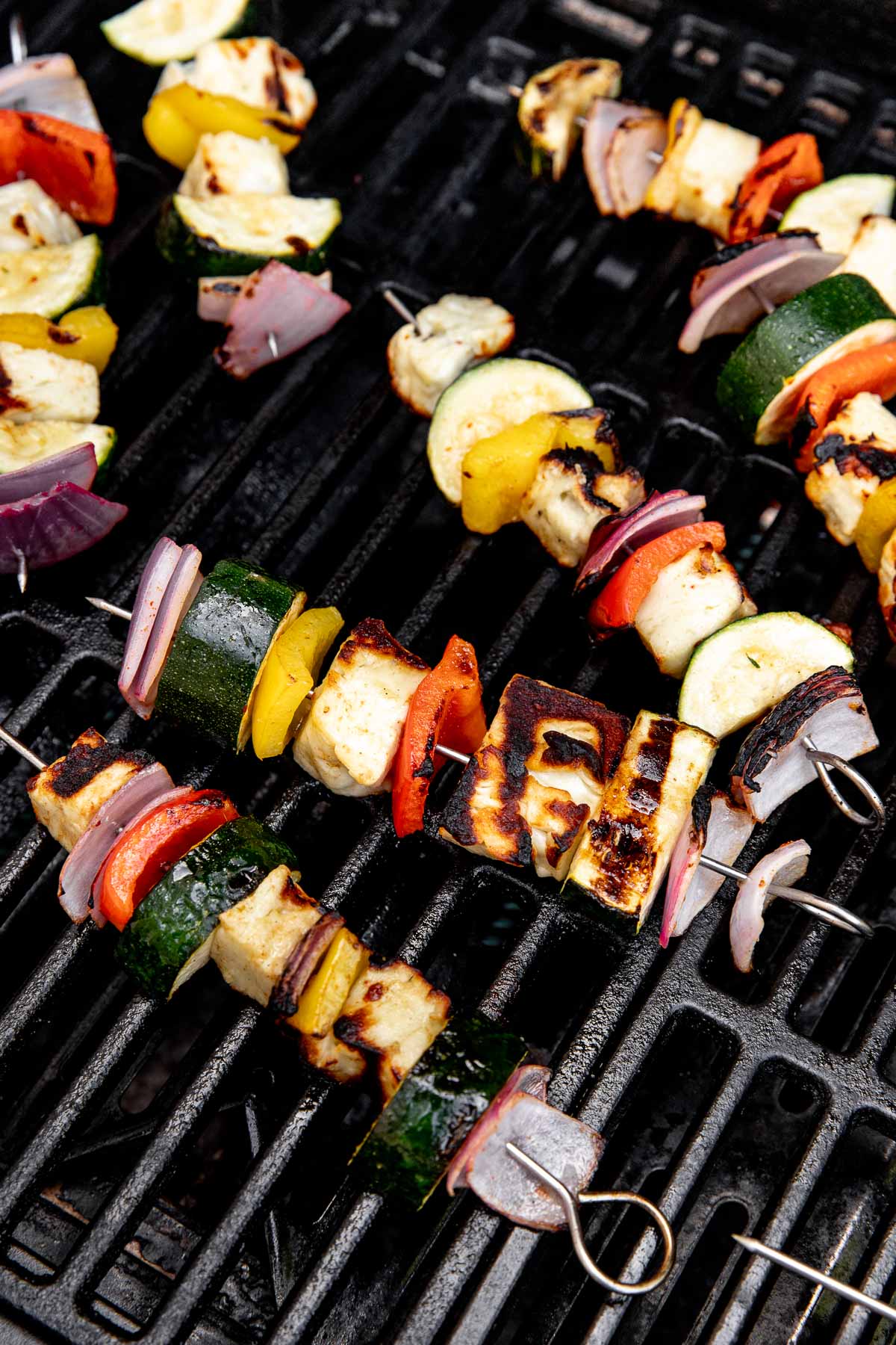 Vegetable skewers with zucchini, bell peppers, red onion, and grilled halloumi cheese cooking on a black grill grate. The veggies and cheese are charred with visible grill marks.