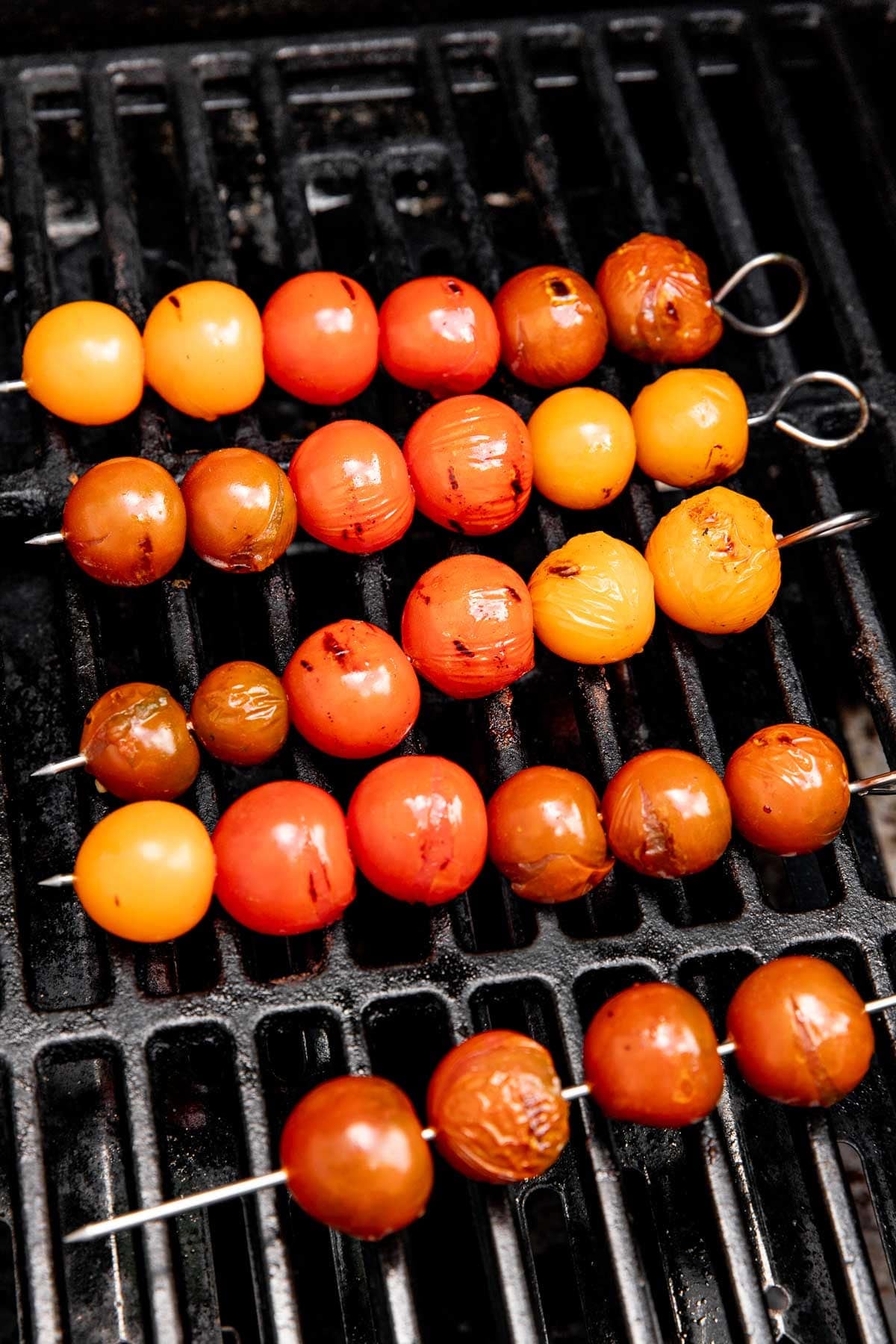 Skewers of assorted colorful cherry tomatoes are grilling on a barbecue, with the tomatoes slightly charred and blistered from the heat. The tomatoes range in shades of red, yellow, and orange.