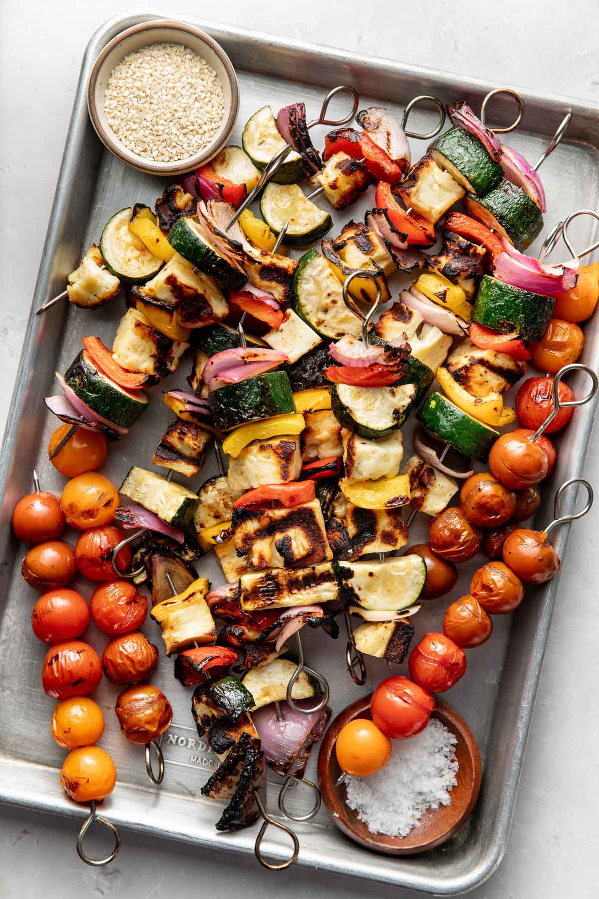 A tray of grilled vegetable skewers with zucchini, red onion, bell pepper, and cherry tomatoes. A small bowl of sesame seeds and a wooden bowl of coarse salt are also on the tray.