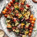 A platter of colorful grilled vegetable skewers with zucchini, cherry tomatoes, red onion, and bell pepper, garnished with fresh herbs and sesame seeds. Small bowls of herbs and salt are nearby.