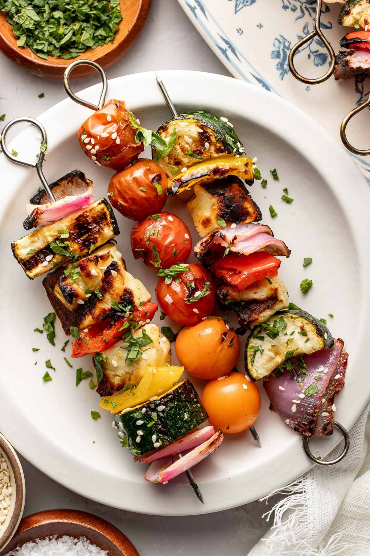 Two skewers on a white plate, each with grilled vegetables and cheese, including cherry tomatoes, zucchini, red onion, bell peppers, and cubes of paneer, garnished with chopped herbs.