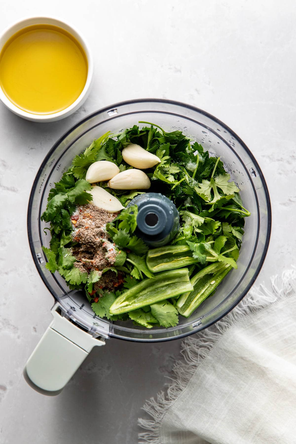 A food processor bowl filled with fresh cilantro, garlic cloves, chopped green chilies, and spices sits on a light countertop next to a small white bowl of olive oil and a fringed cloth napkin.