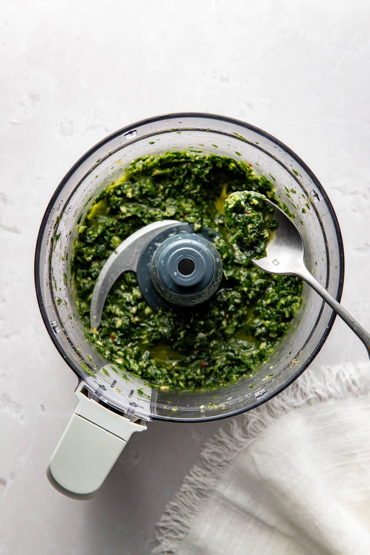 A food processor containing a freshly blended green herb mixture, with a metal spoon scooping some out. The processor sits on a light surface next to a white cloth.