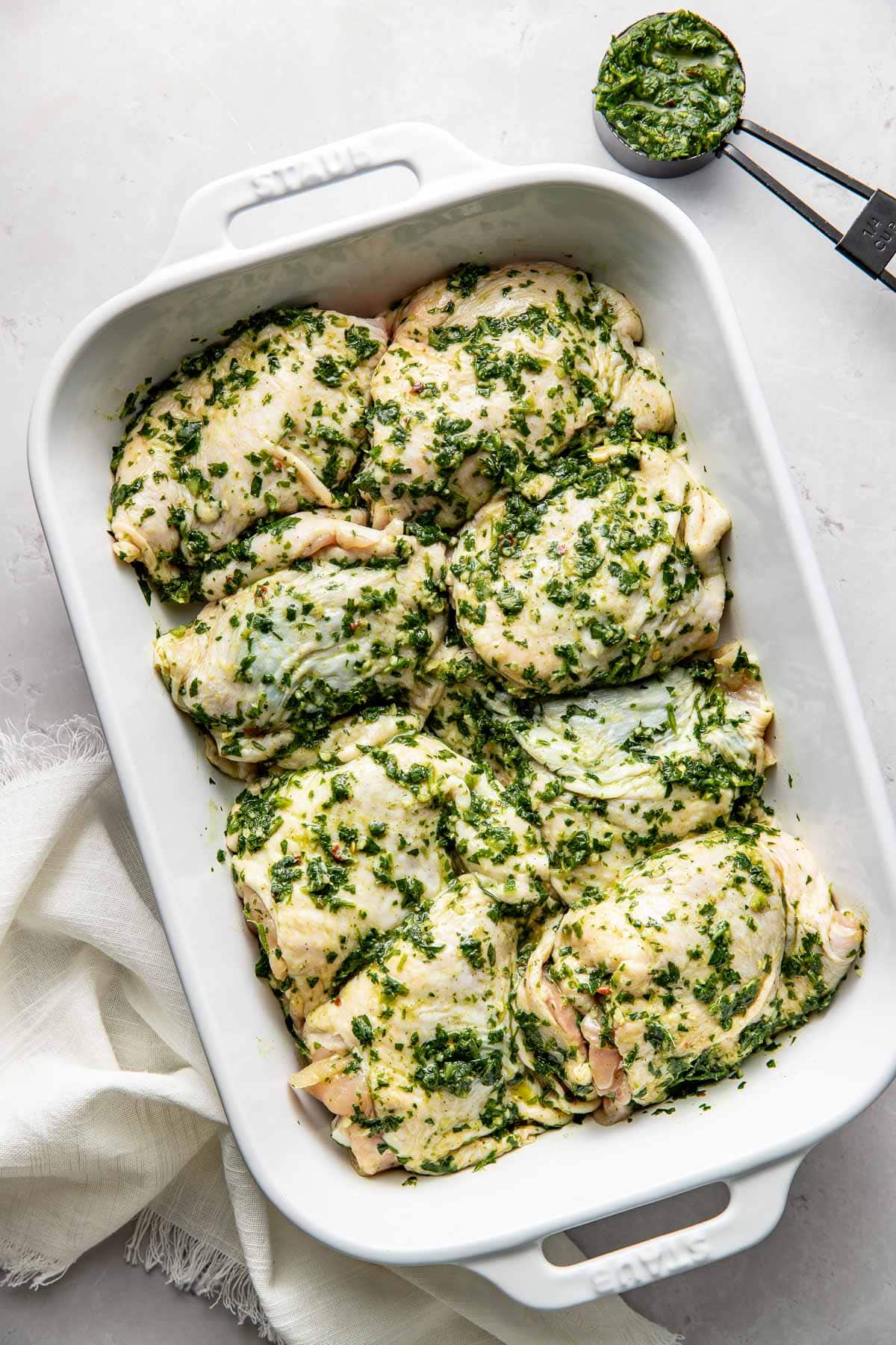Raw chicken pieces coated in a green herb marinade are arranged in a white baking dish. A small cup of additional marinade with measuring spoons is nearby on a light surface. A white cloth sits beside the dish.