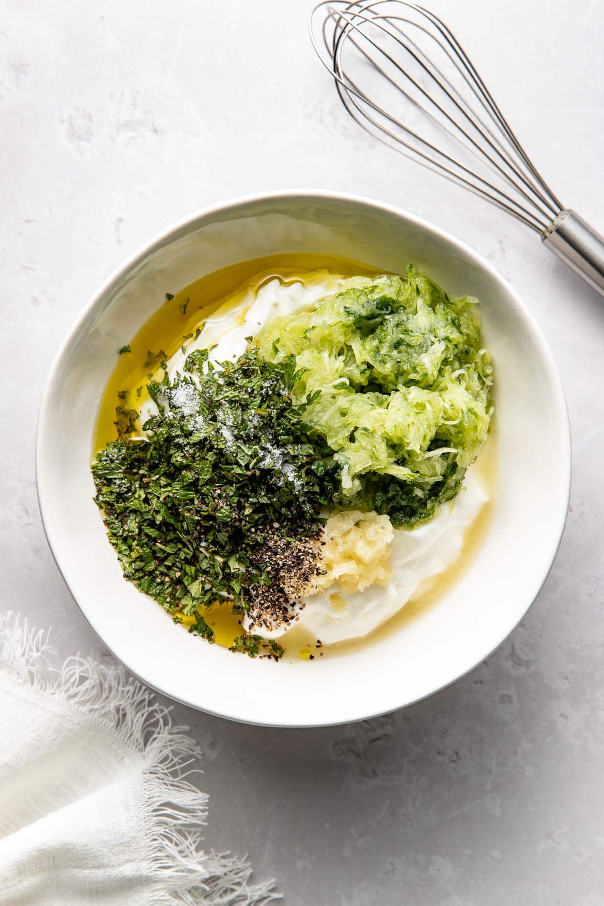A bowl containing yogurt, shredded cucumber, chopped herbs, minced garlic, ground black pepper, olive oil, and salt, with a whisk placed beside the bowl on a light-colored surface.