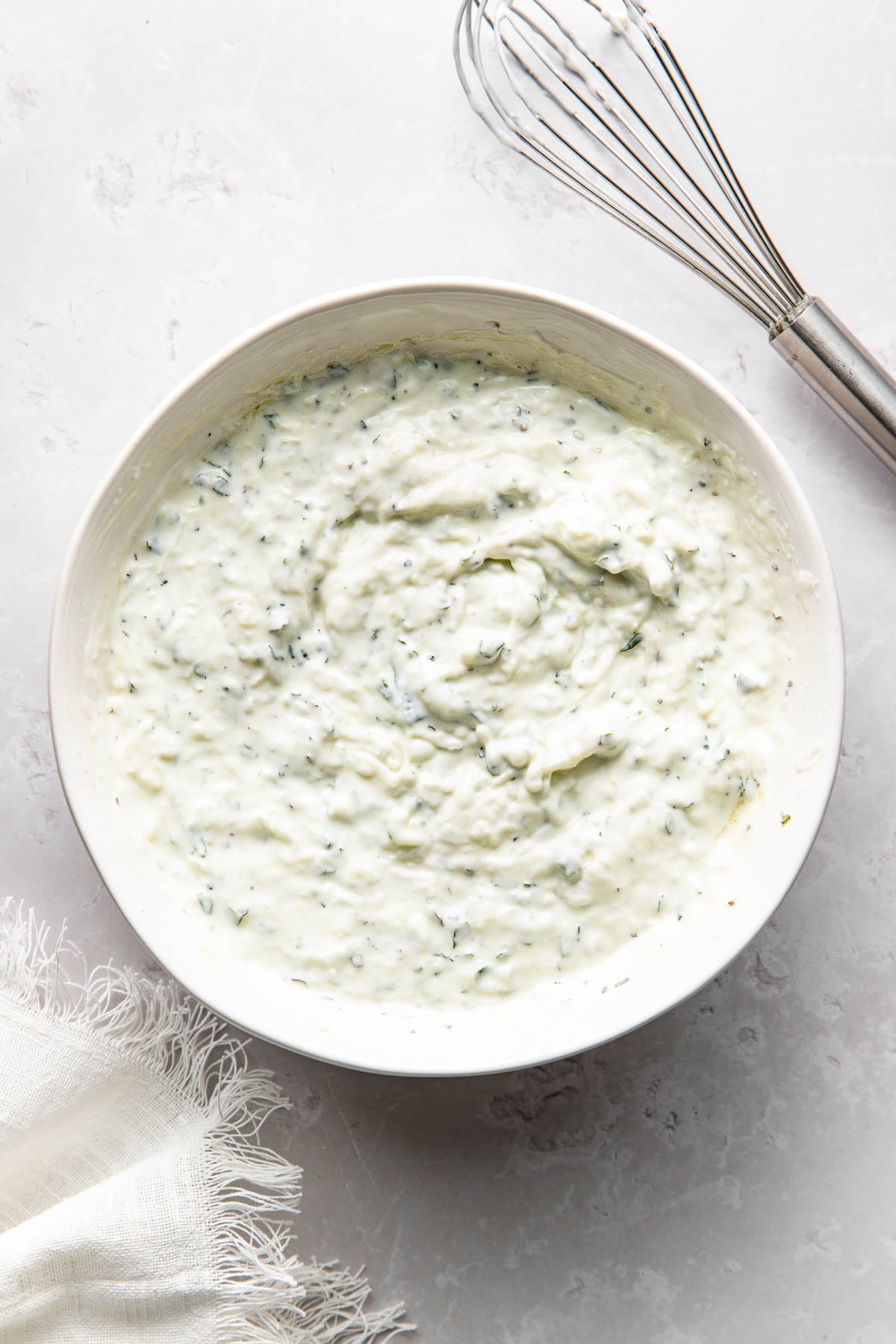 A white bowl filled with creamy tzatziki sauce garnished with herbs sits on a light gray surface next to a metal whisk and a white cloth napkin.