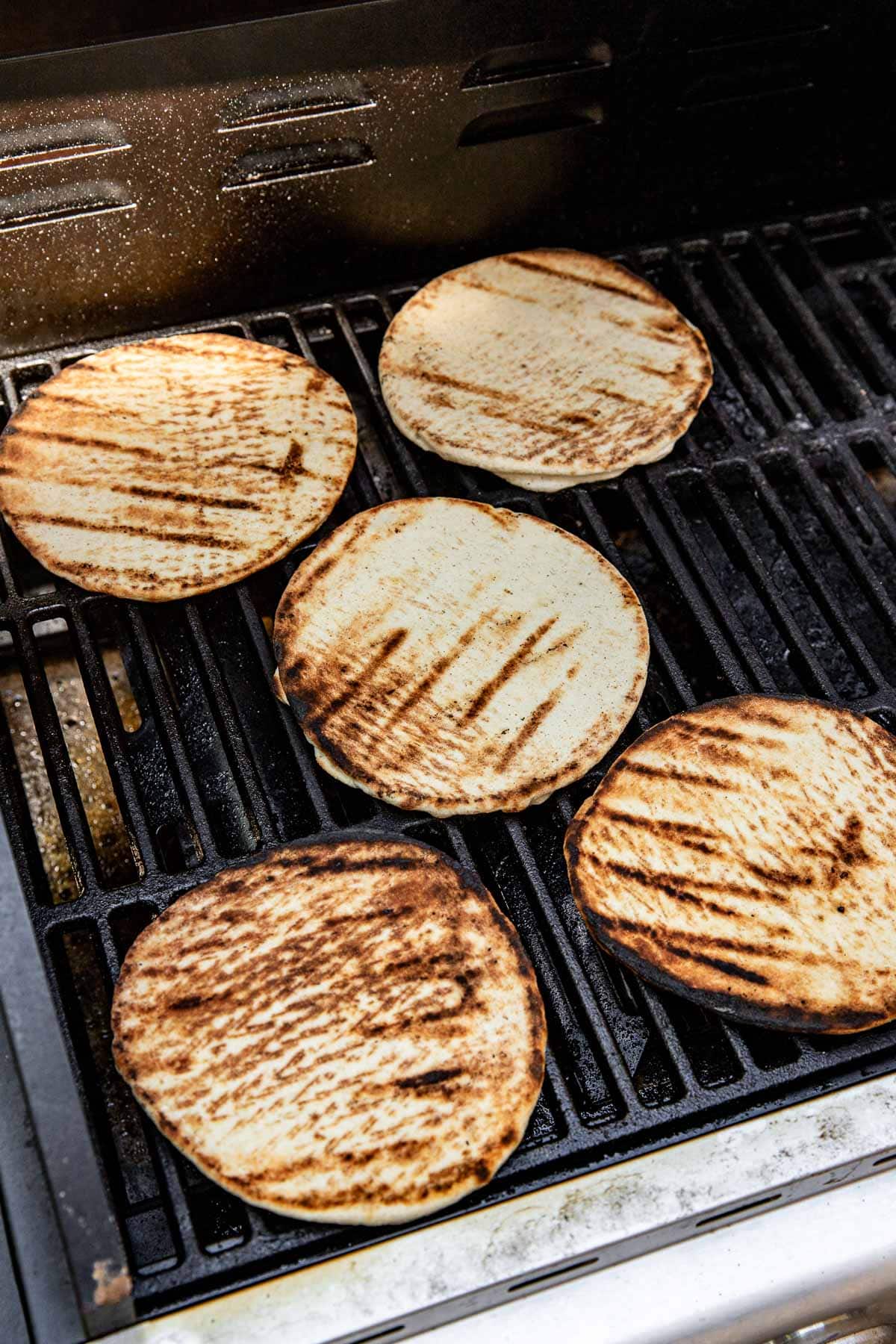 Five round pieces of flatbread with grill marks are cooking on an outdoor grill.