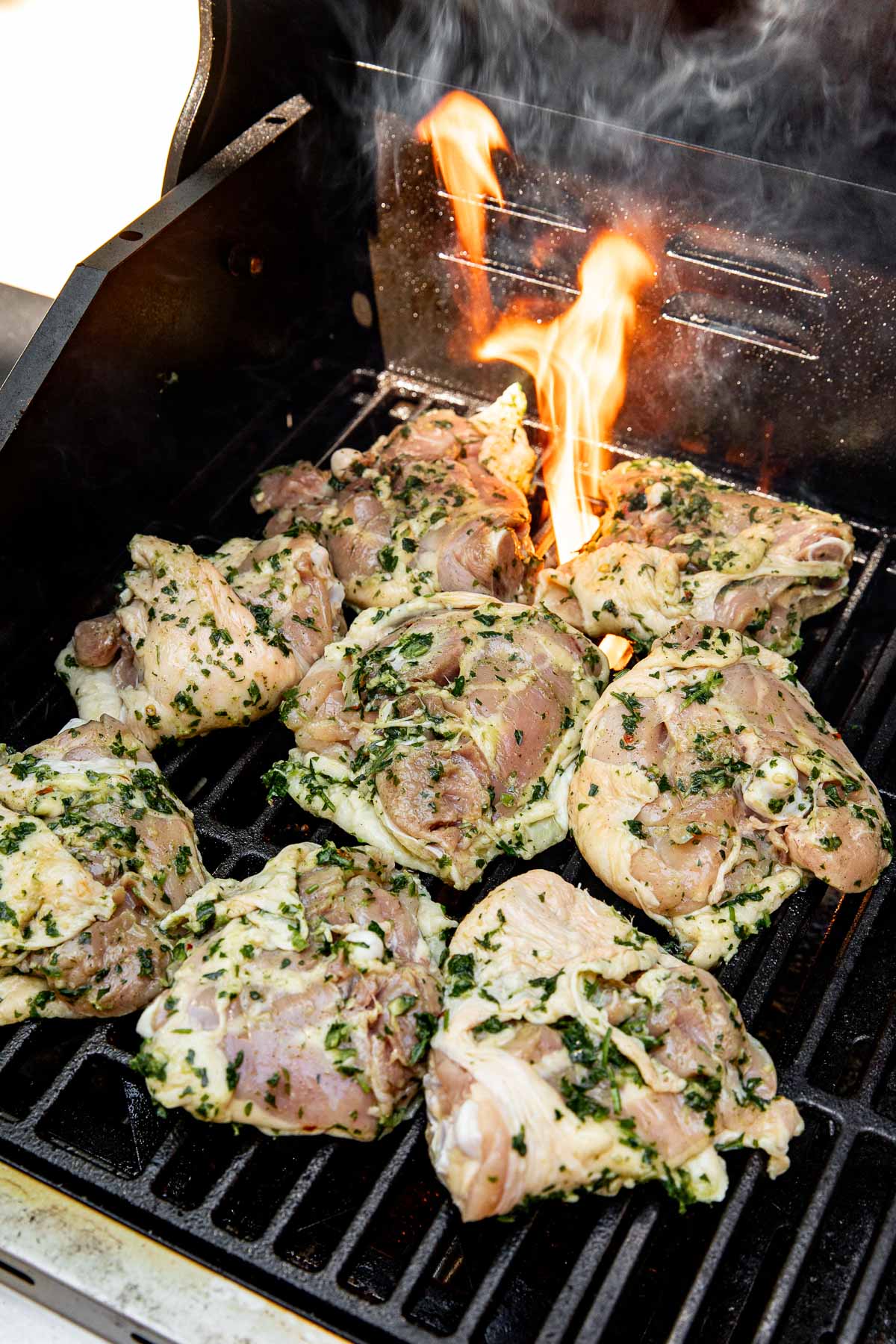 Seasoned chicken thighs with herbs and garlic are grilling on an open barbecue, with flames rising in the background and smoke visible, indicating active cooking.
