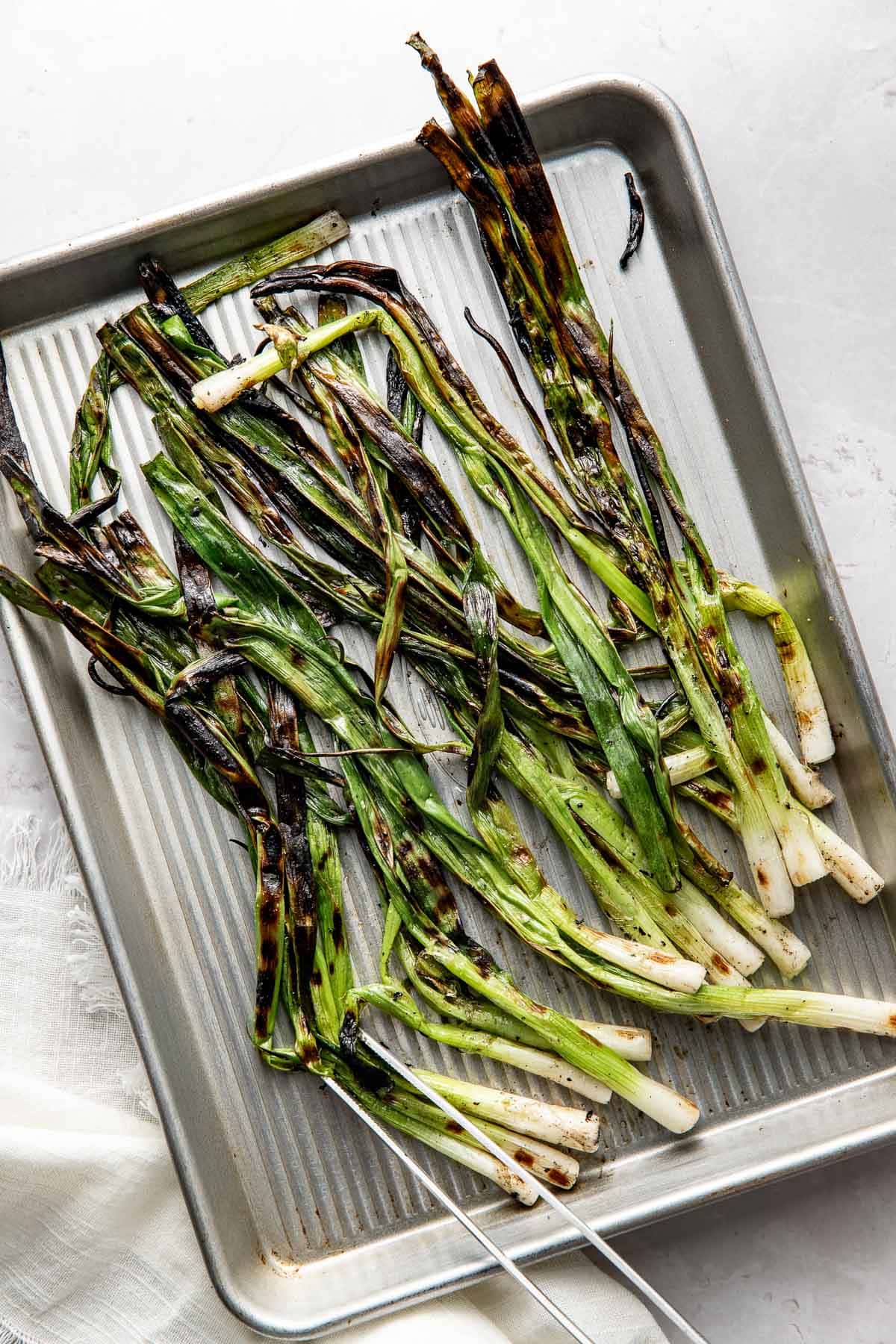 Grilled scallions with charred edges arranged on a metal baking sheet, displaying a mix of green and browned colors. The baking sheet rests on a white surface with part of a cloth visible in the corner.