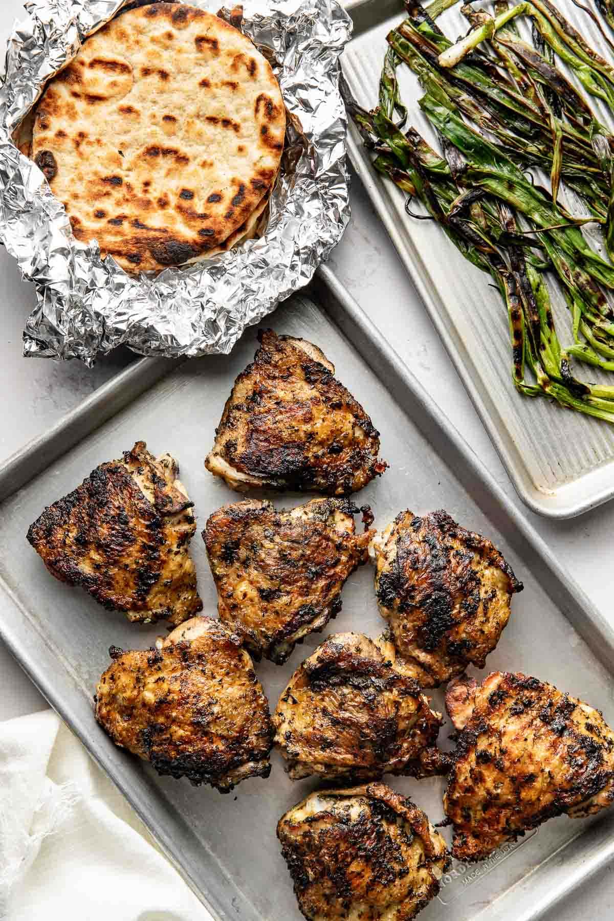 A baking tray with seven grilled chicken thighs, a foil-wrapped stack of flatbreads, and a plate of grilled green onions arranged on a light surface.