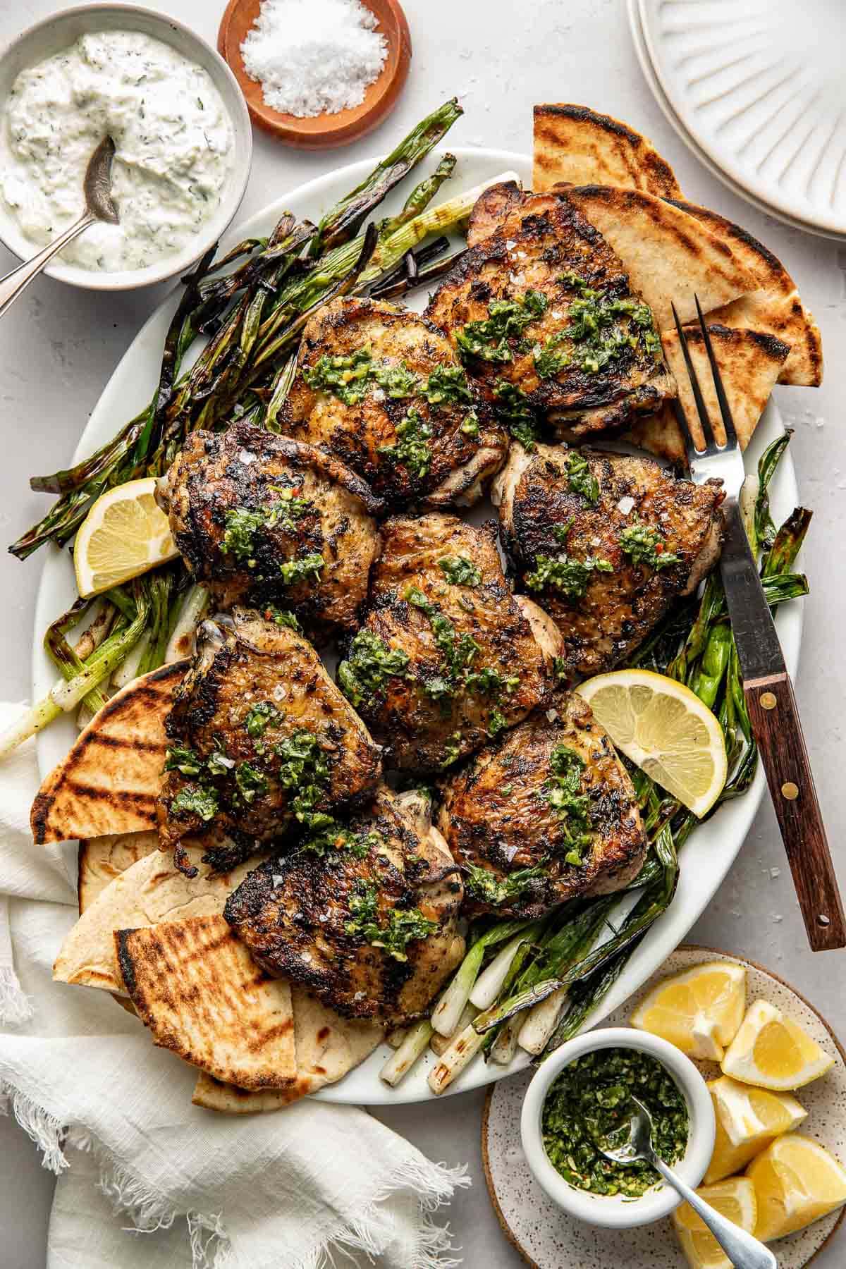 A platter of grilled chicken thighs garnished with herbs, served with grilled pita bread, asparagus, lemon wedges, and a side of tzatziki sauce. Fork, knife, and small bowls are arranged around the plate.