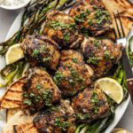 A platter with grilled chicken thighs garnished with herbs, surrounded by grilled pita bread, charred green onions, lemon wedges, and small bowls of sauces on the side.
