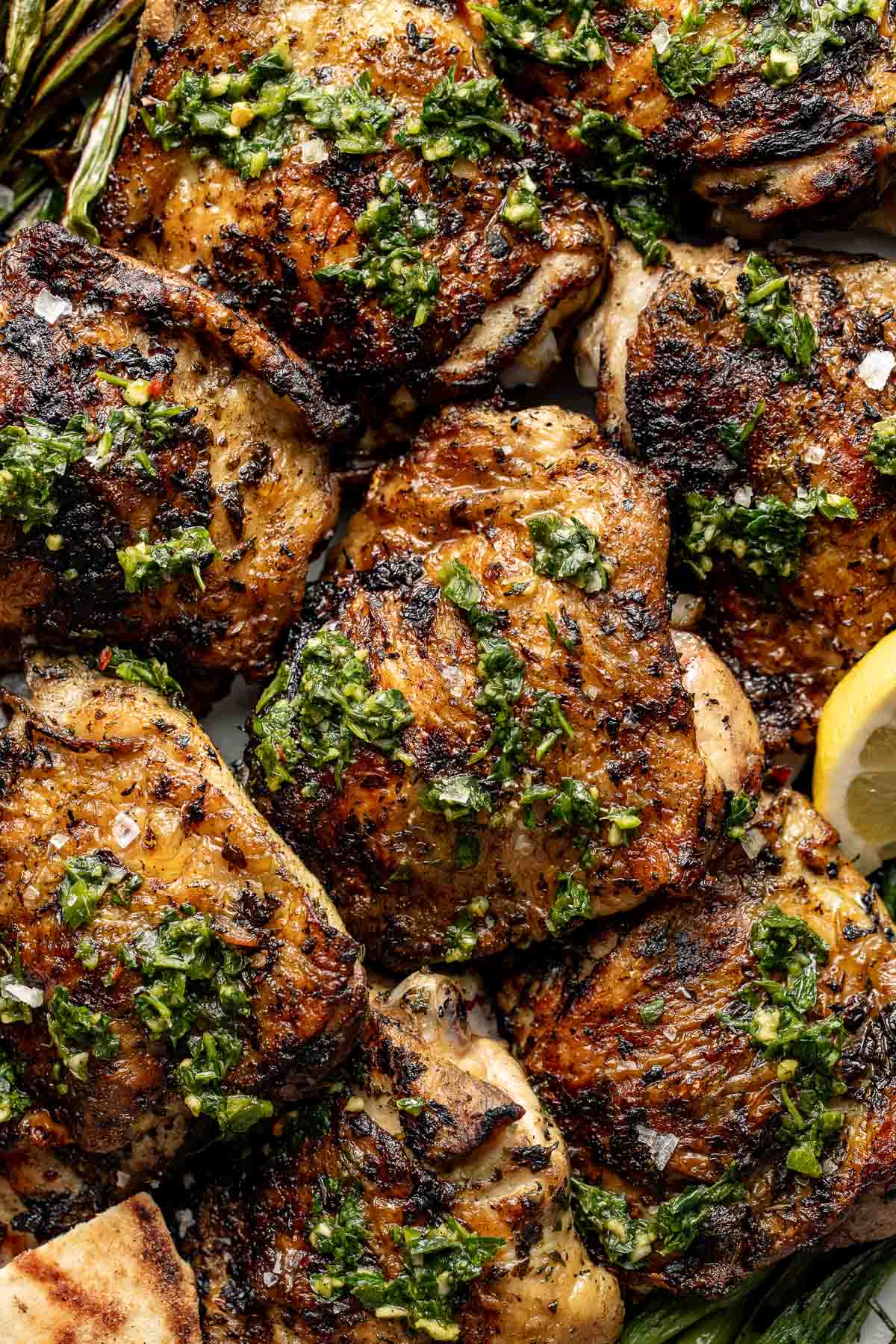 Grilled chicken thighs topped with fresh green chimichurri sauce, garnished with herbs and served with lemon wedges. The chicken has a charred, crispy appearance.