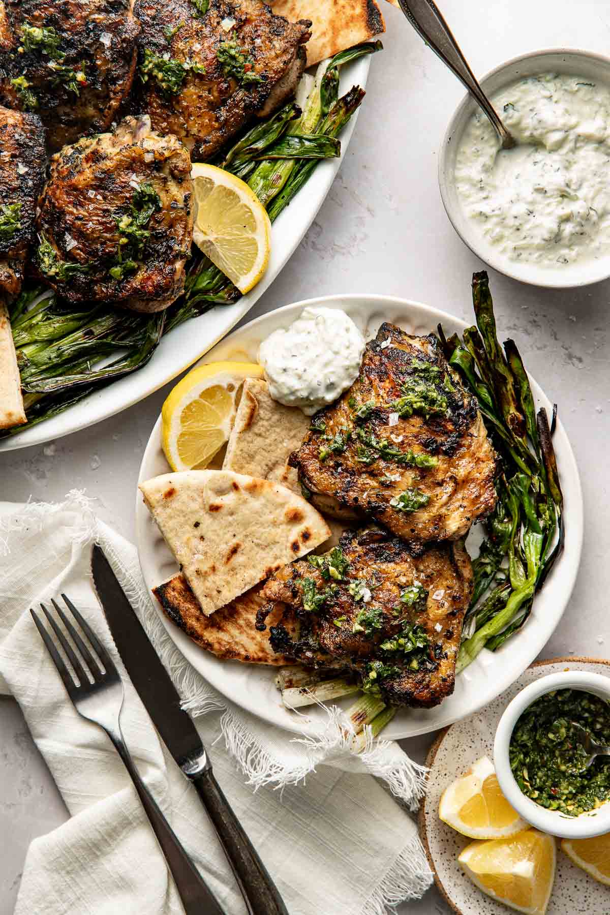 A plate with grilled chicken, flatbread, grilled green onions, lemon slices, and a dollop of tzatziki sauce. A fork and knife are placed on a napkin beside the plate, with extra sauce, lemons, and food in the background.