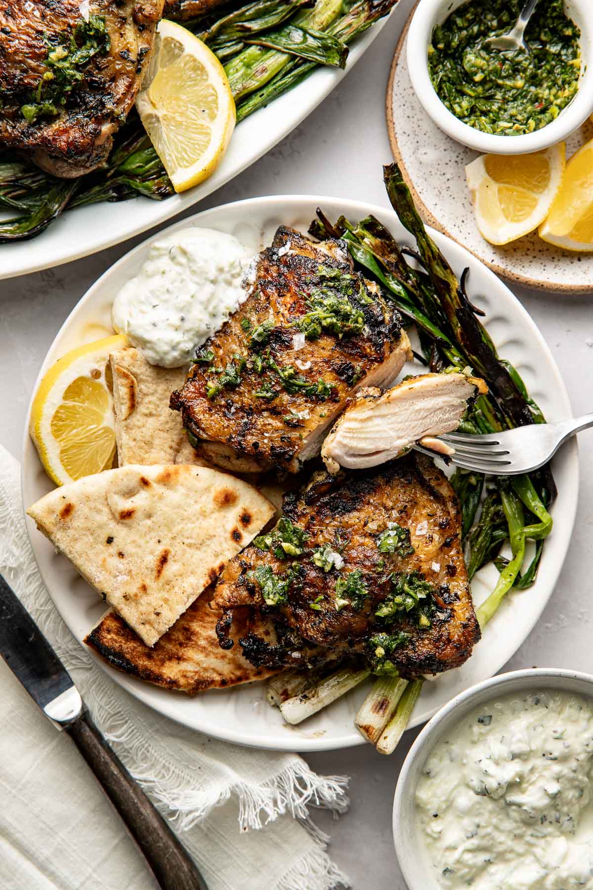 A plate with grilled chicken thighs topped with green herb sauce, grilled scallions, pita bread, lemon wedges, and two dollops of creamy white sauce. A fork holds a piece of chicken. Another bowl of sauce and lemon slices are nearby.