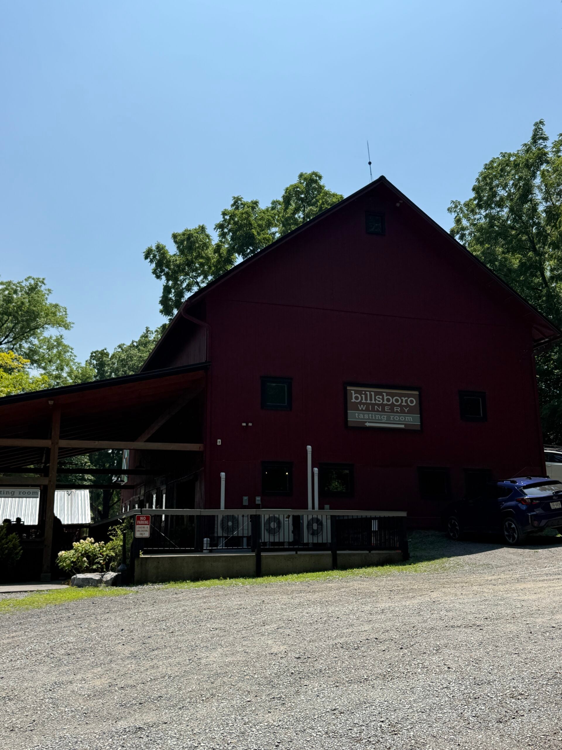 A large red barn with a sign reading billsboro winery stands beside a gravel lot, surrounded by trees under a clear blue sky.