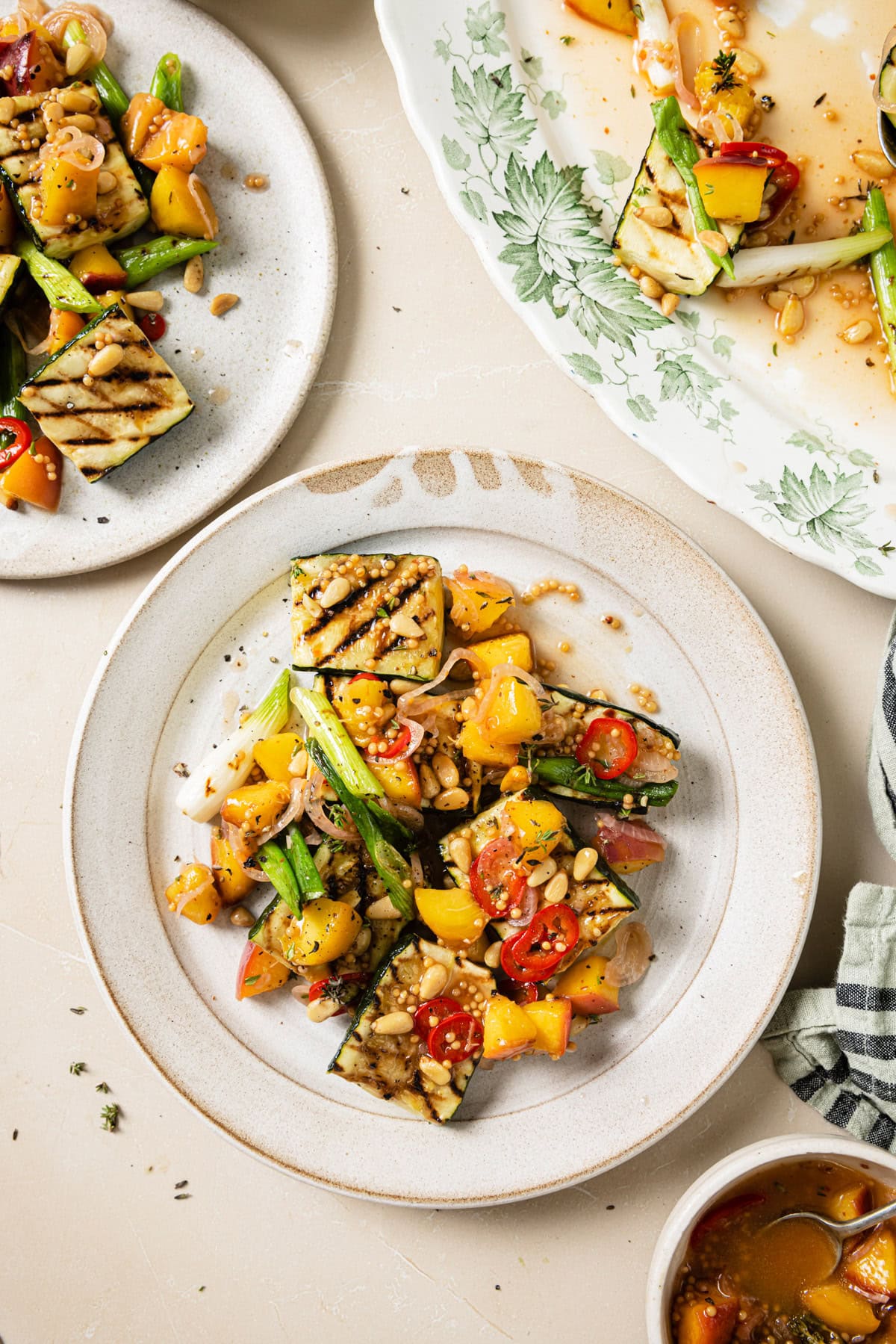 A plate of grilled zucchini topped with chopped yellow and red tomatoes, green beans, pine nuts, and herbs, with more vegetables and sauce on nearby plates and dishware.