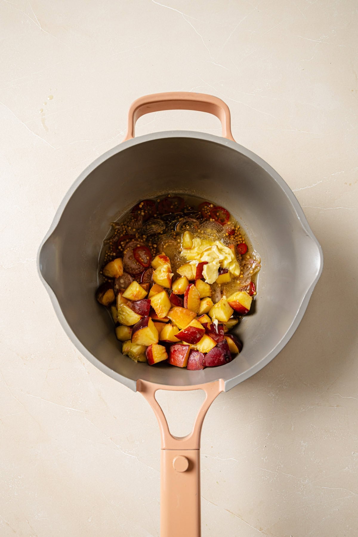 Overhead view of a saucepan containing chopped plums, butter, and sugar on a light countertop. The ingredients appear to be starting to cook and mix together.