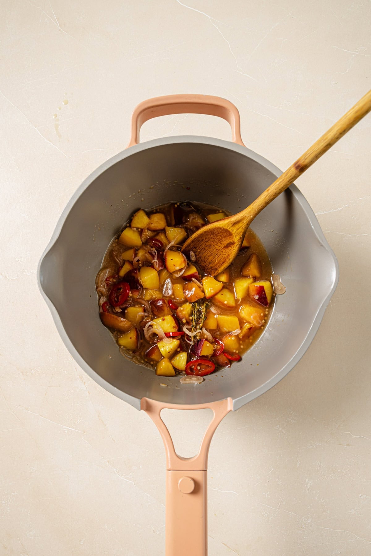 A saucepan with a wooden spoon, containing a simmering mixture of chopped peaches, red chili slices, and herbs in liquid, sits on a light countertop.