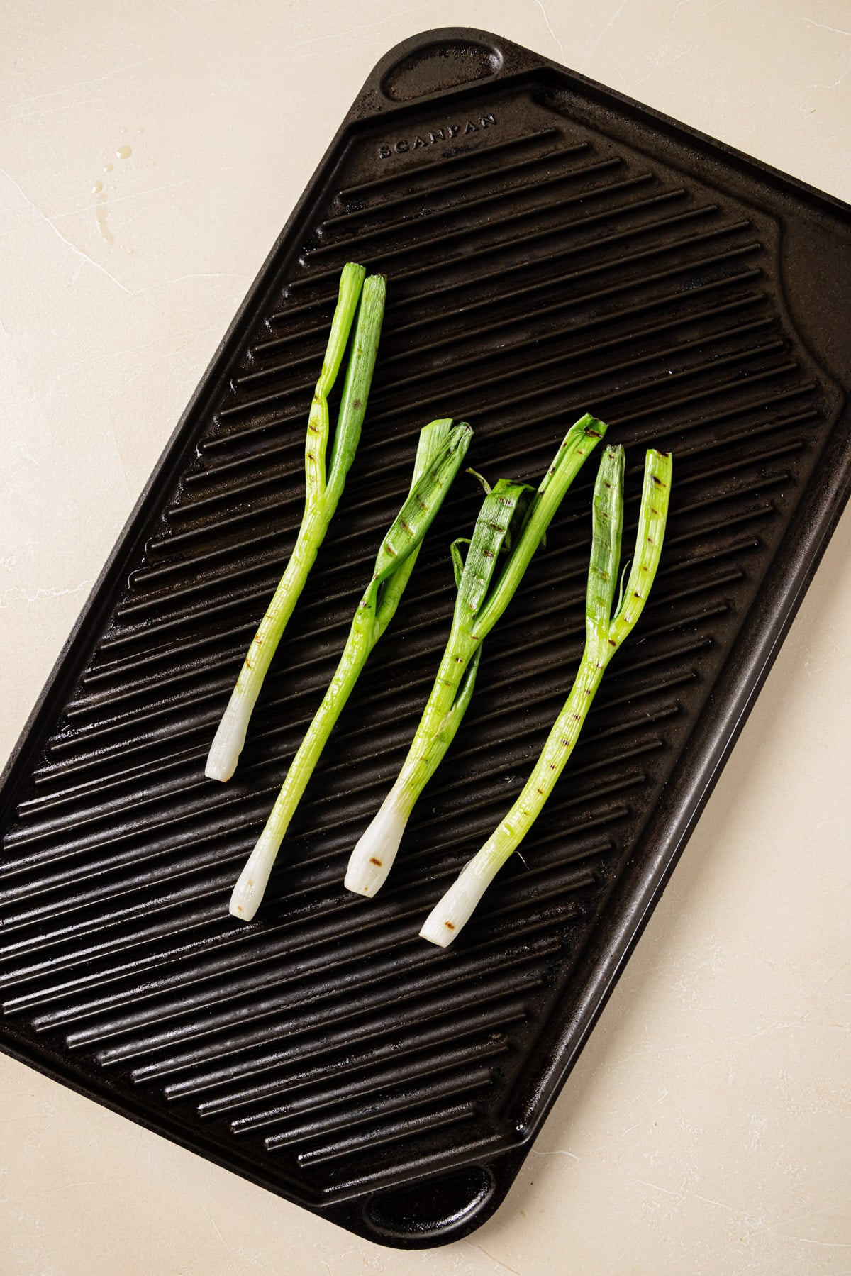 Four green onions are arranged in a row on a black ridged grill pan set on a light-colored surface.