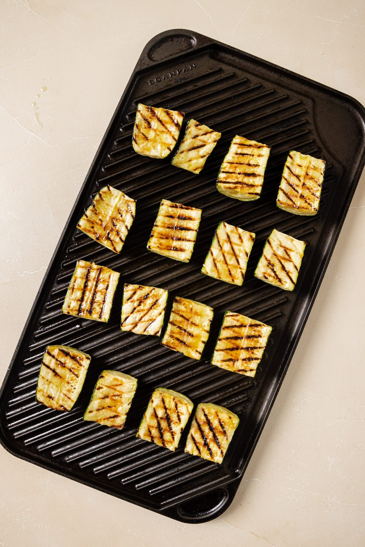 Rectangular slices of zucchini with grill marks are arranged on a black grill pan, which sits on a light-colored surface.