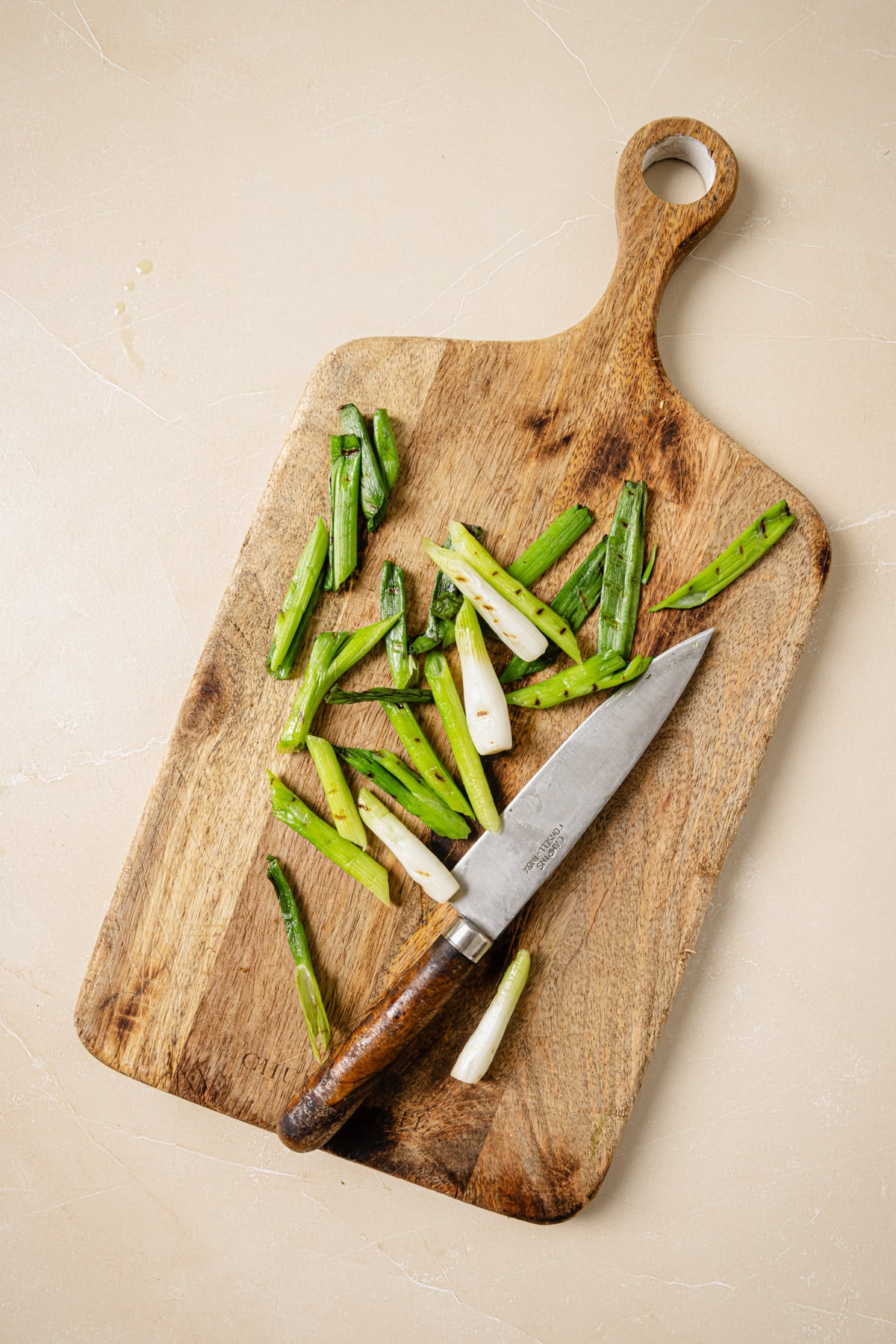 Sliced green onions and a knife rest on a wooden cutting board with a handle, placed on a light beige surface.