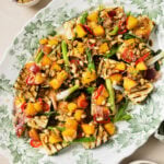 A platter of grilled vegetables topped with diced mango, tomatoes, pine nuts, herbs, and a drizzle of dressing. Small bowls of pine nuts and dressing are nearby on a beige surface.