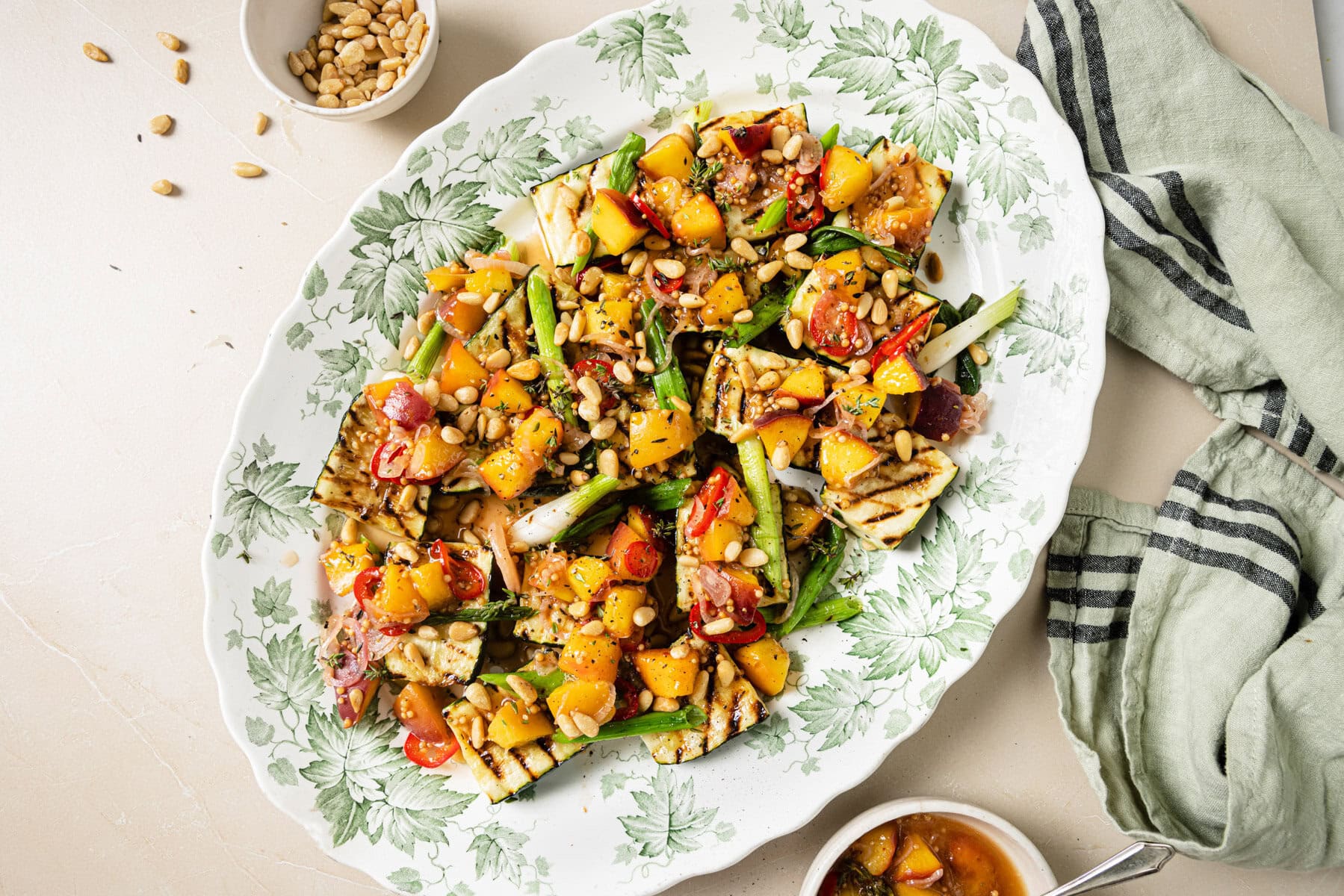 A platter of grilled halloumi salad with pineapple, tomatoes, spring onions, red chili, and pine nuts, served on a leafy-patterned dish with a small bowl of pine nuts and a green striped cloth nearby.