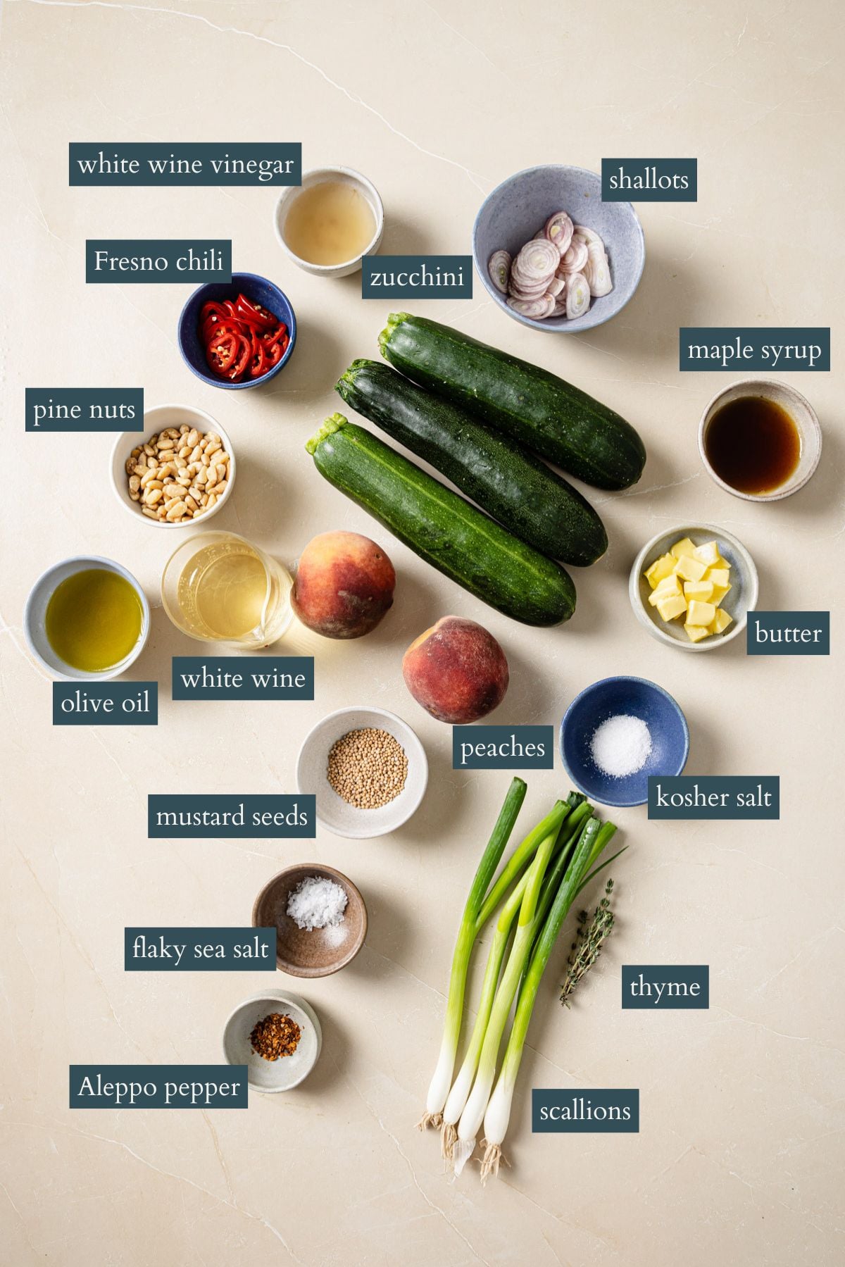 Labeled ingredients on a beige surface: zucchini, peaches, Fresno chili, pine nuts, olive oil, white wine, mustard seeds, Aleppo pepper, scallions, thyme, flaky sea salt, kosher salt, maple syrup, butter, shallots, white wine vinegar.