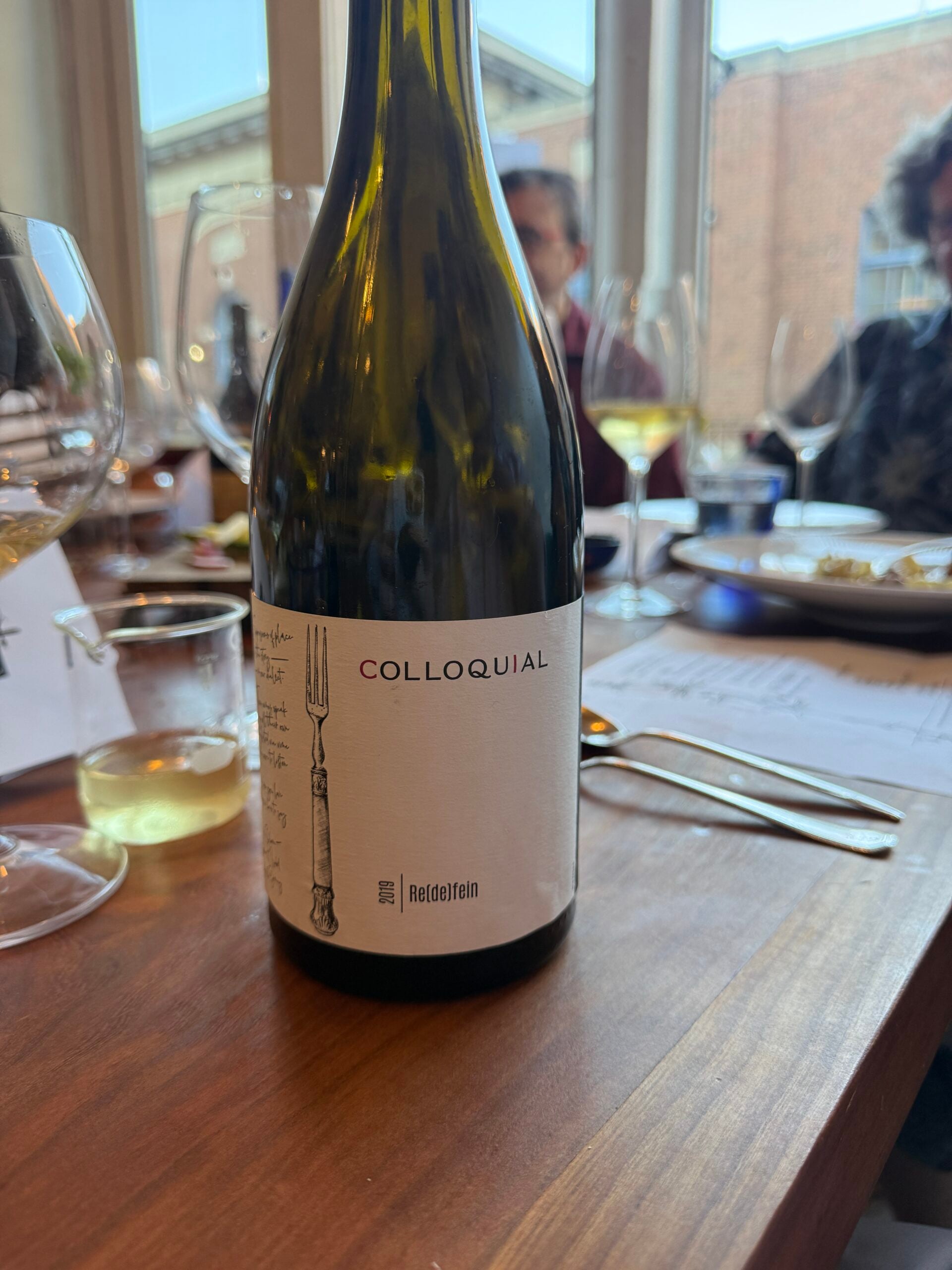 A close-up of a wine bottle labeled “Colloquial” on a wooden table, surrounded by wine glasses, paper, and blurred people in the background.
