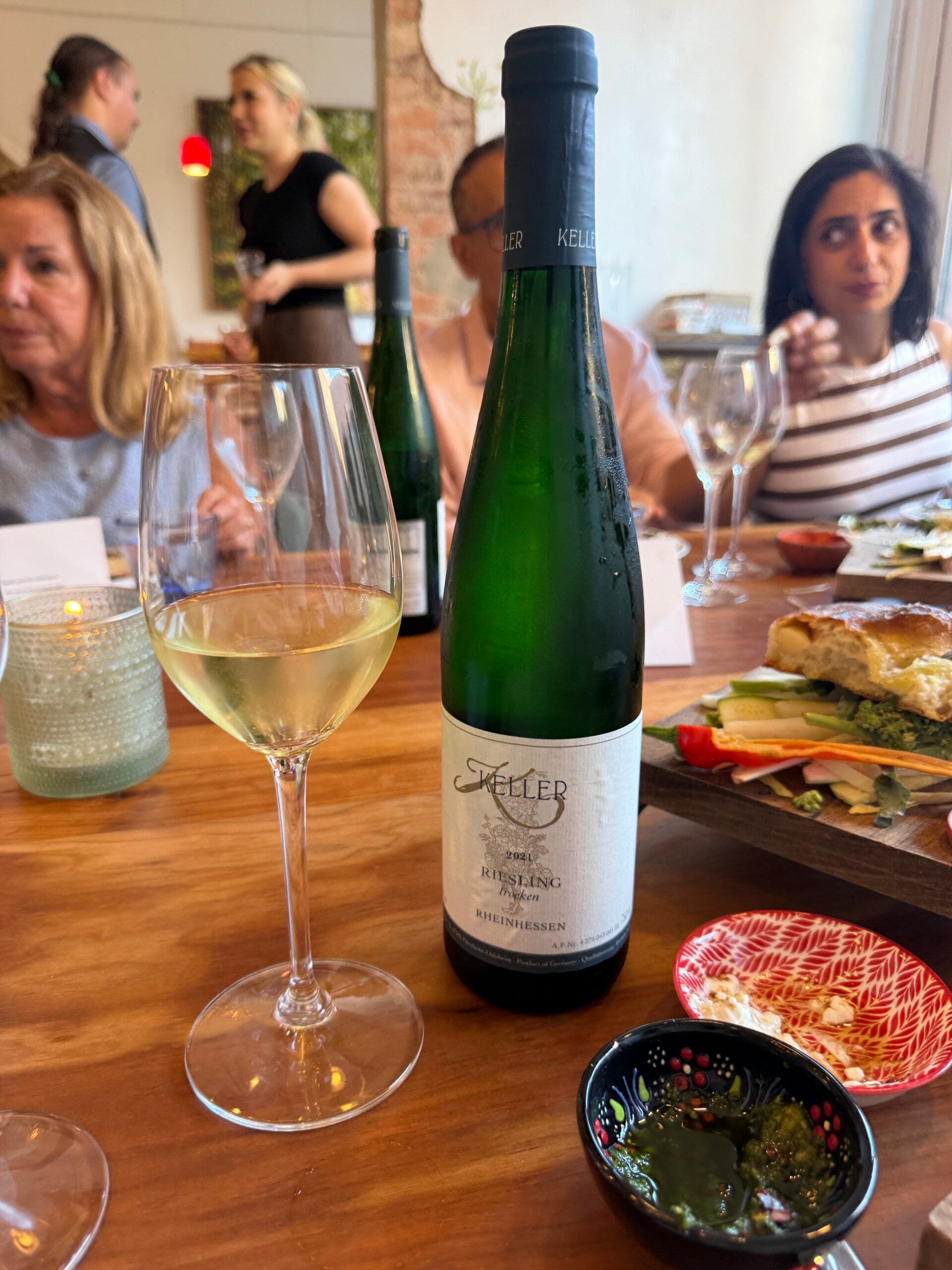 A bottle of Keller Riesling wine and a glass of white wine on a wooden table with food, surrounded by people dining in a restaurant.